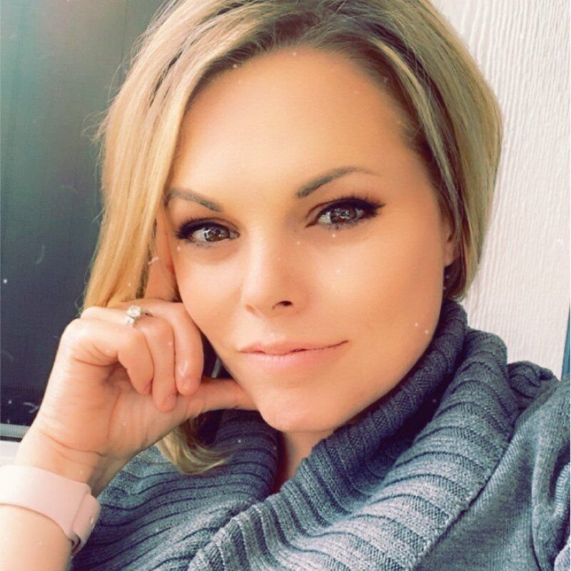 A woman with blonde hair and blue eyes, smiling softly, wearing a gray sweater and a pink watch.