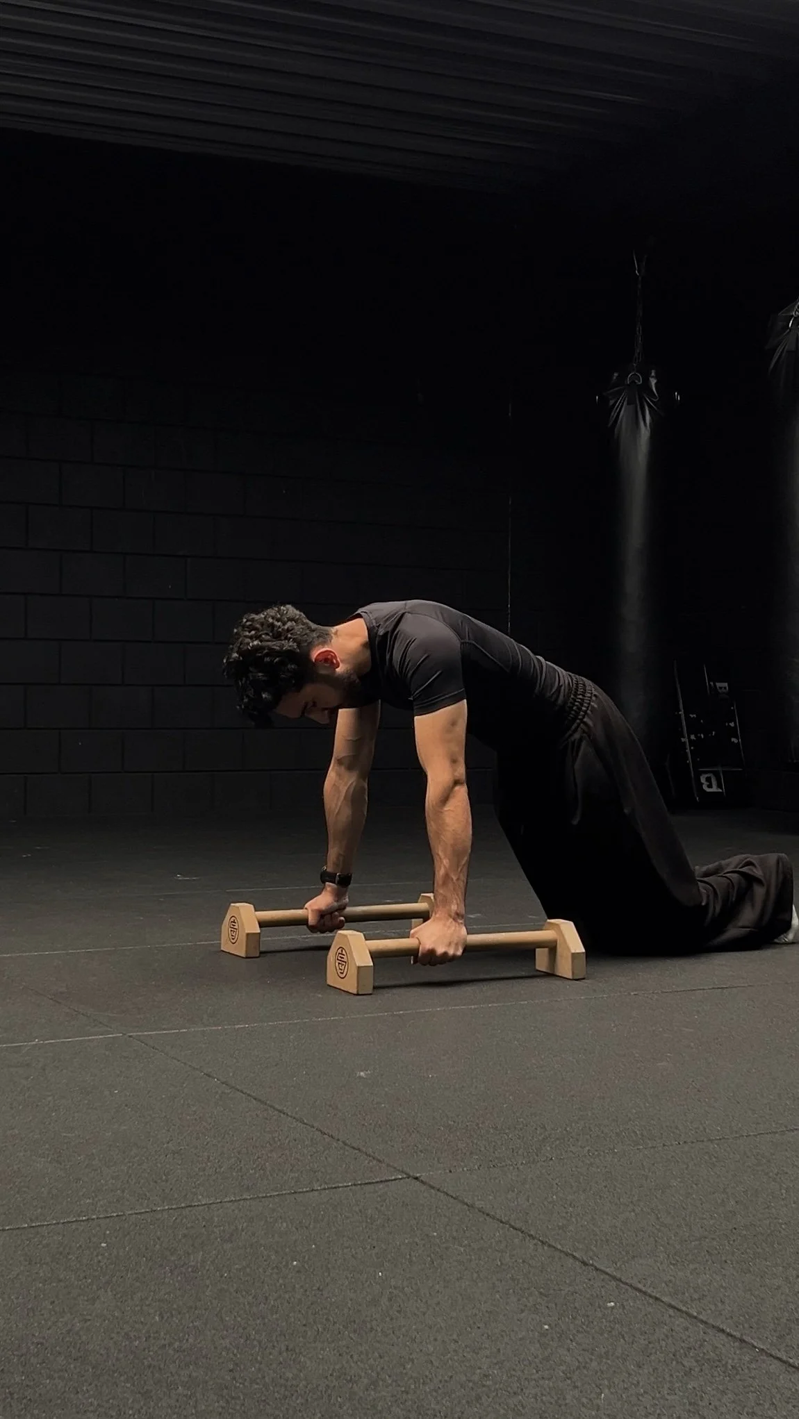 A man performing a forearm plank exercise on a wooden push-up aid on a black gym floor, with black walls and gym equipment in the background.