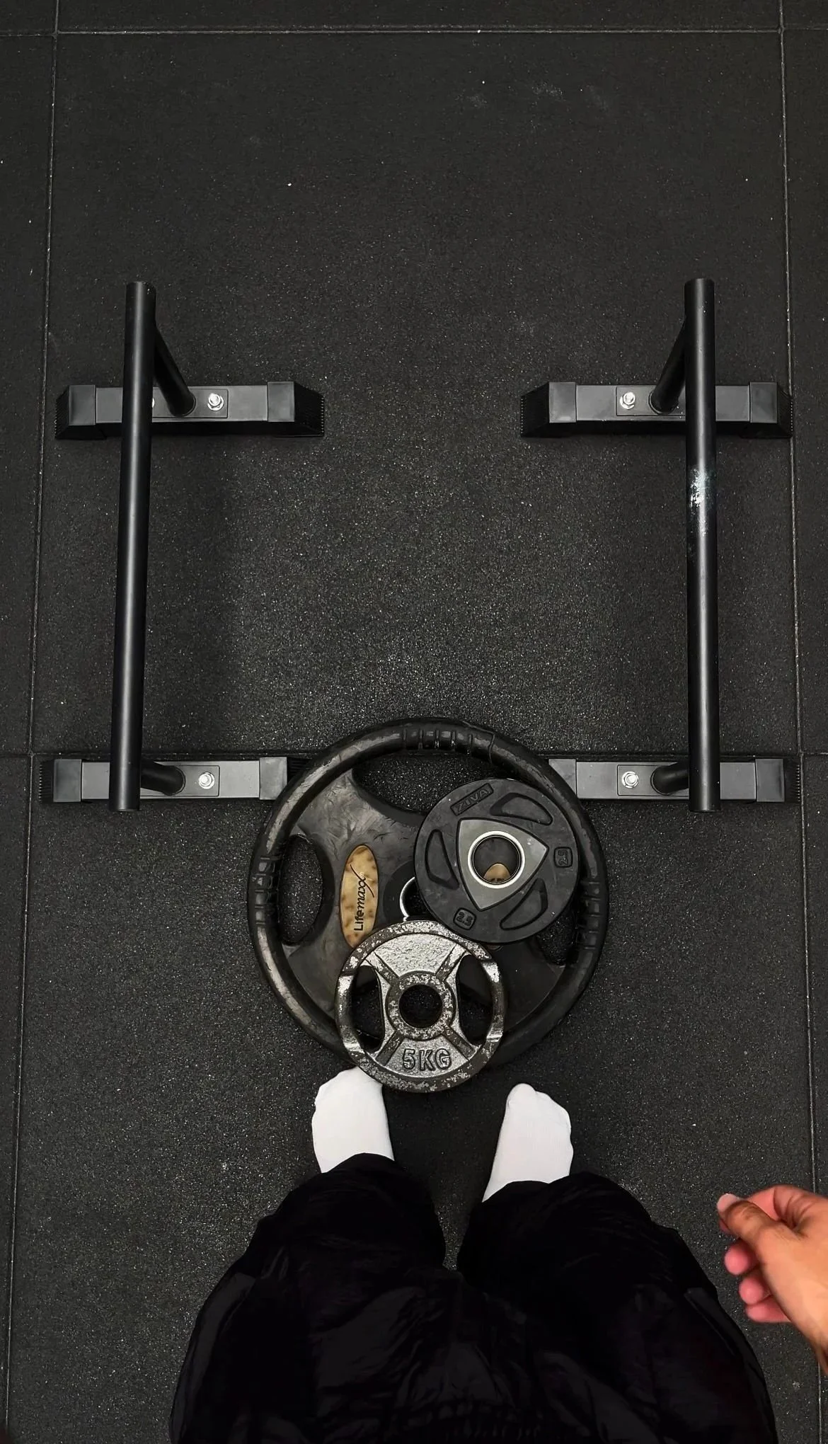View from above of a person standing with feet on a gym floor with a weight plate and parallettes in front.