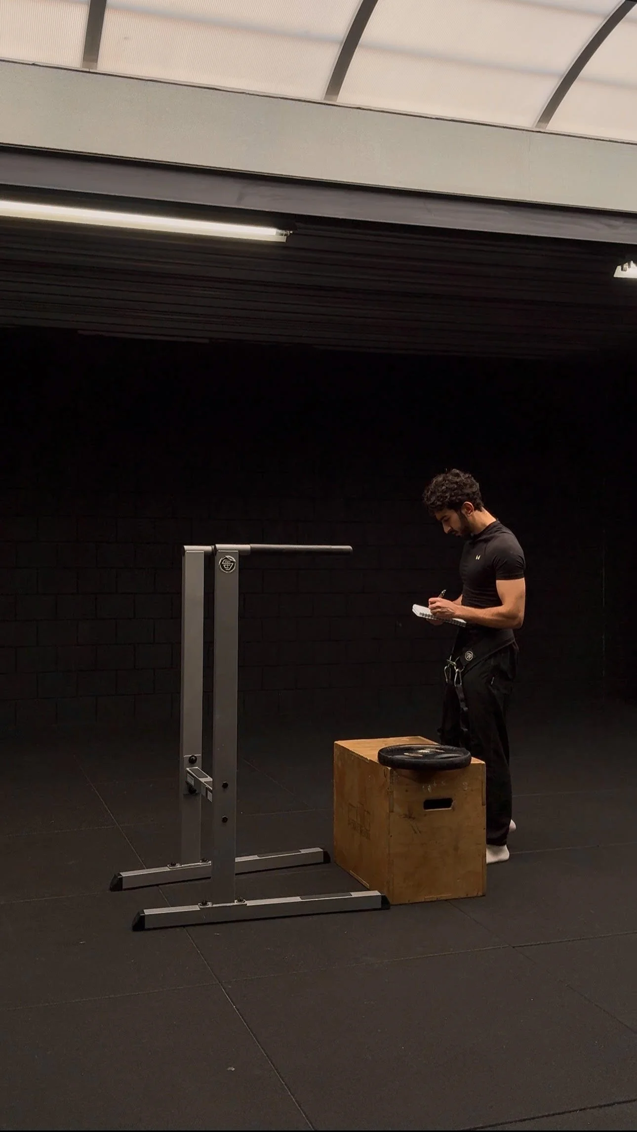 A man in a training area writing on a notepad next to a wooden plyometric box and a Dip station.