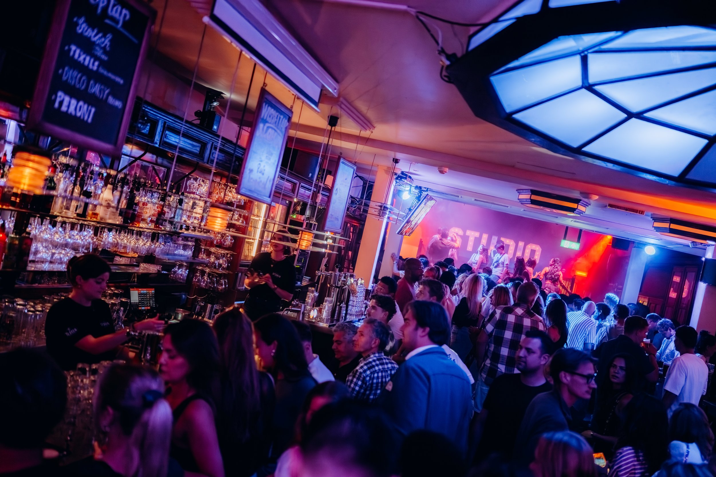 People dancing and socializing inside a vibrant nightclub with a bar and stage lighting.