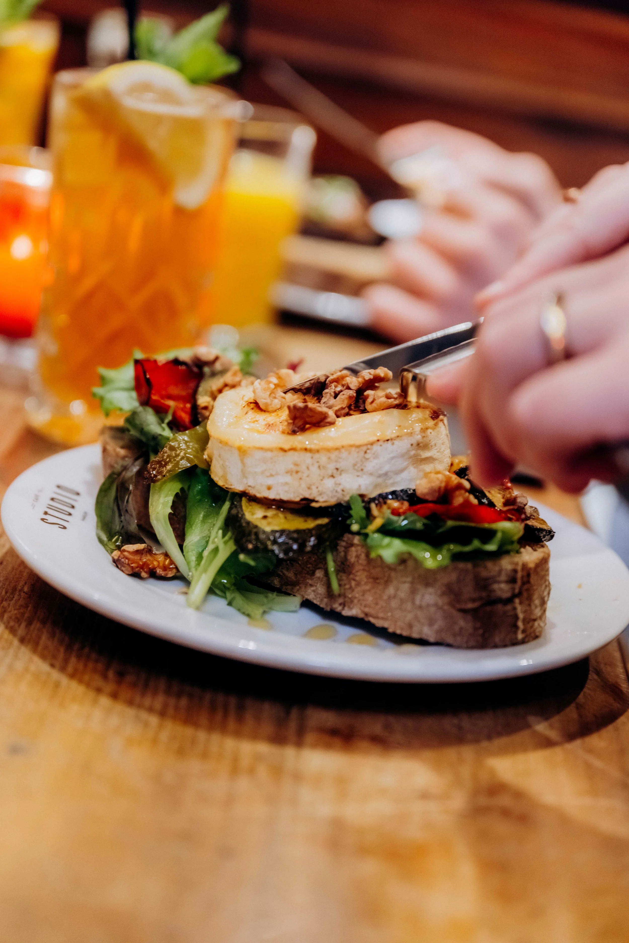 A person using tongs to pick up a slice of cake topped with whipped cream and nuts, served on a plate with salad and grilled vegetables, with drinks in the background.