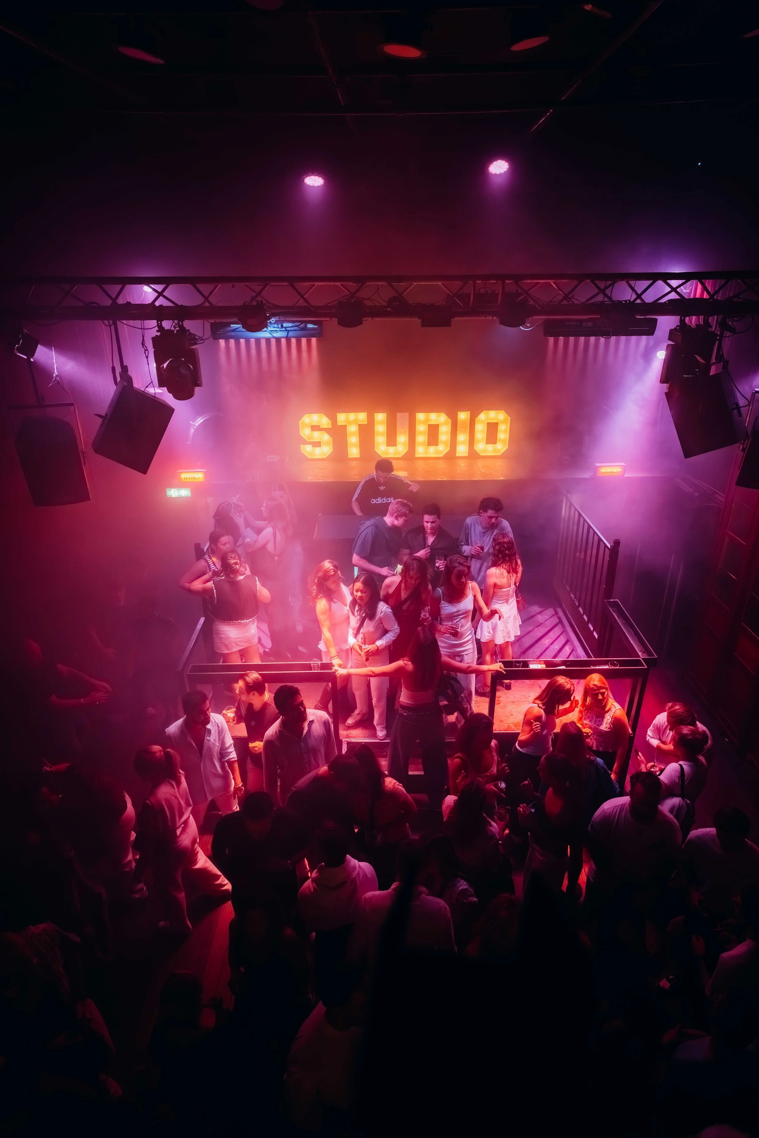 People dancing and socializing at a nightclub or concert venue with colorful lighting and a stage overhead that reads 'STUDIO' in bright lights.