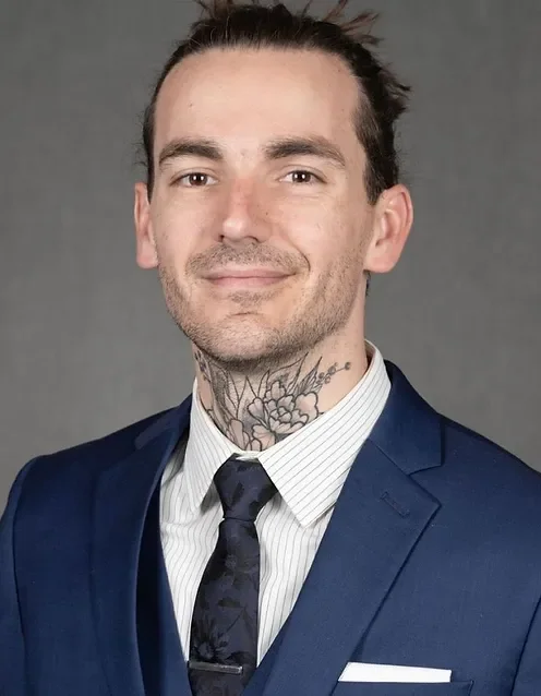 A man in a blue suit, white dress shirt, and dark tie, with tattoos on his neck, smiling against a gray background.
