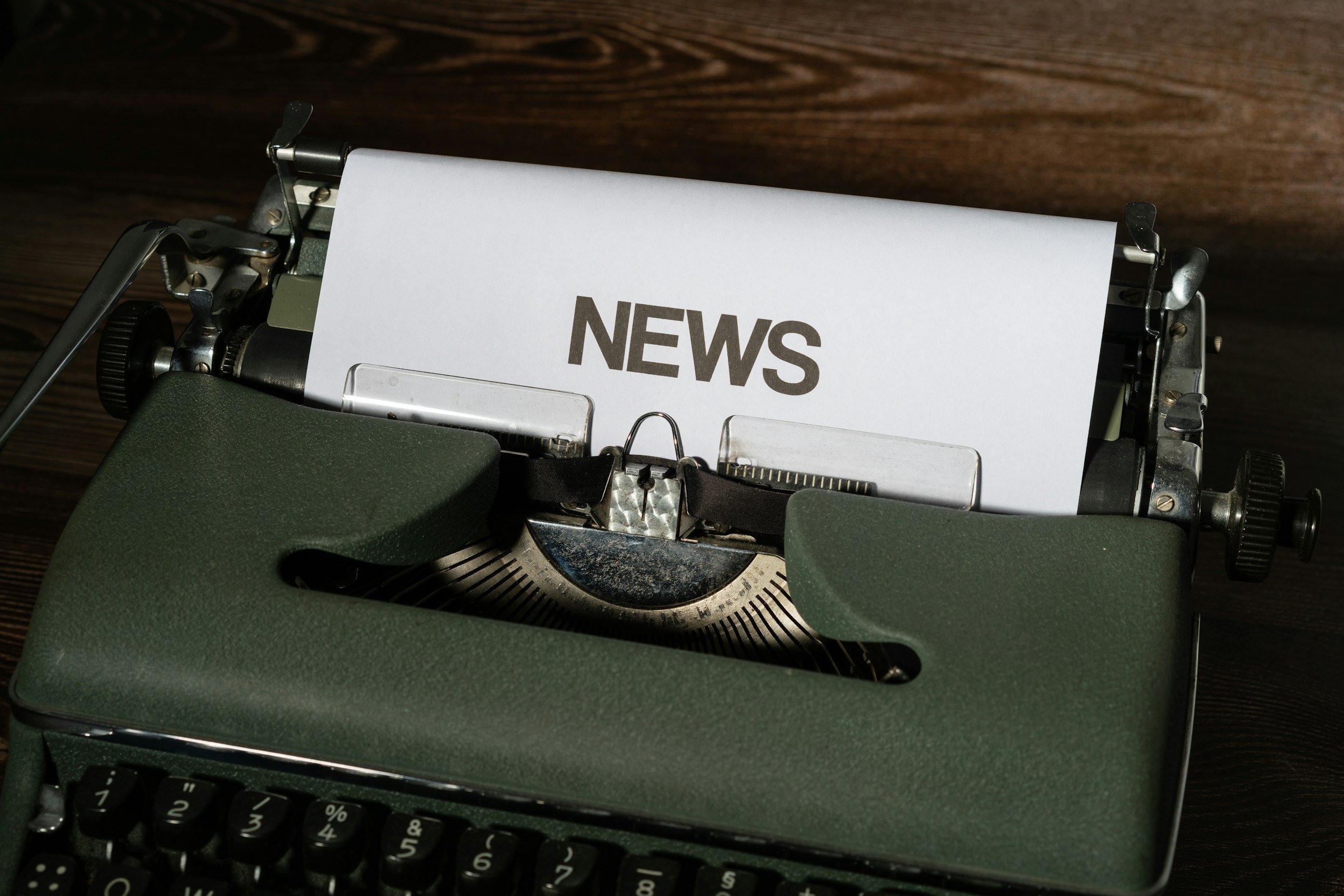 A vintage green typewriter with a paper that says 'NEWS' in bold letters, placed on a wooden surface.