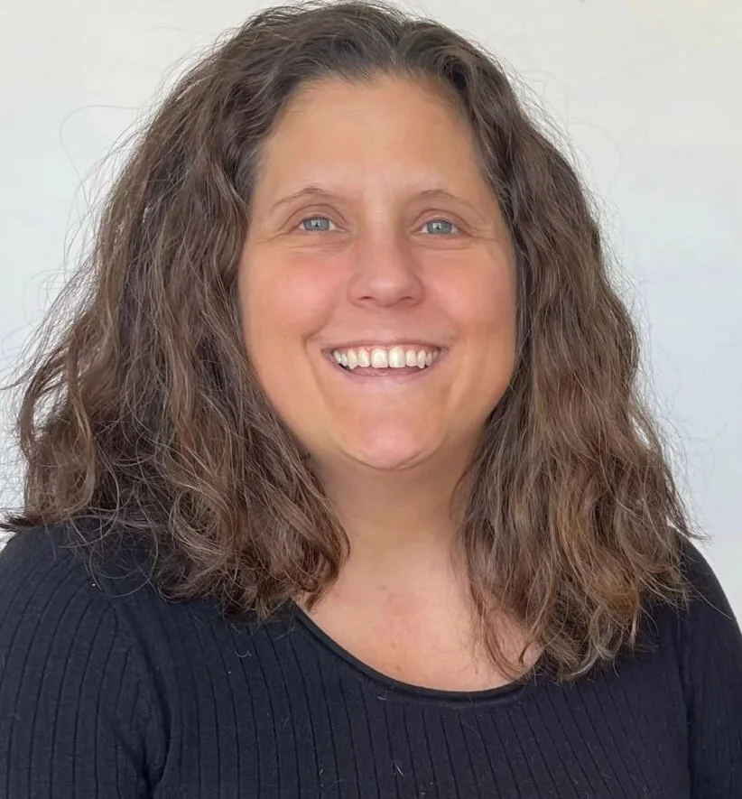 A woman with wavy brown hair and blue eyes, smiling and wearing a black ribbed top, against a plain background.