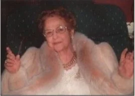An elderly woman with short curly hair and glasses, sitting on a chair, smiling, wearing a white top and a beige fur coat.