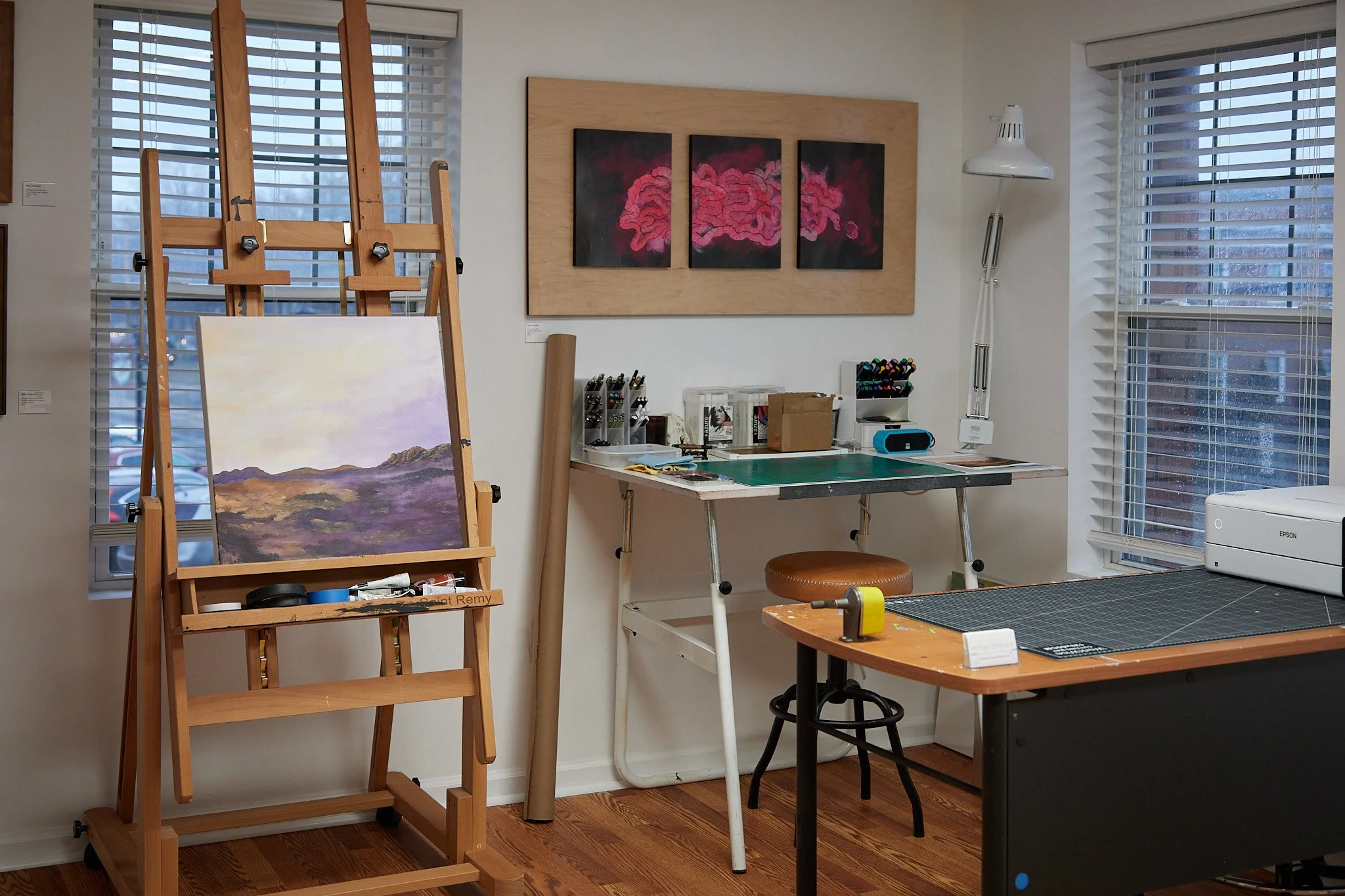 An art studio with an easel holding a landscape painting, a table with art supplies, a brown stool, and two large windows with blinds letting in natural light.