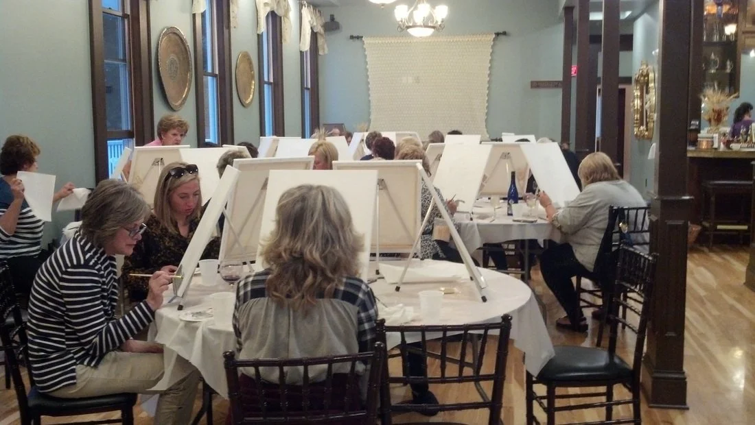 A group of people participating in a painting class in a cozy, decorated room with blue walls, large windows, and a chandelier. They are sitting around tables with canvases on easels, focused on their artwork.