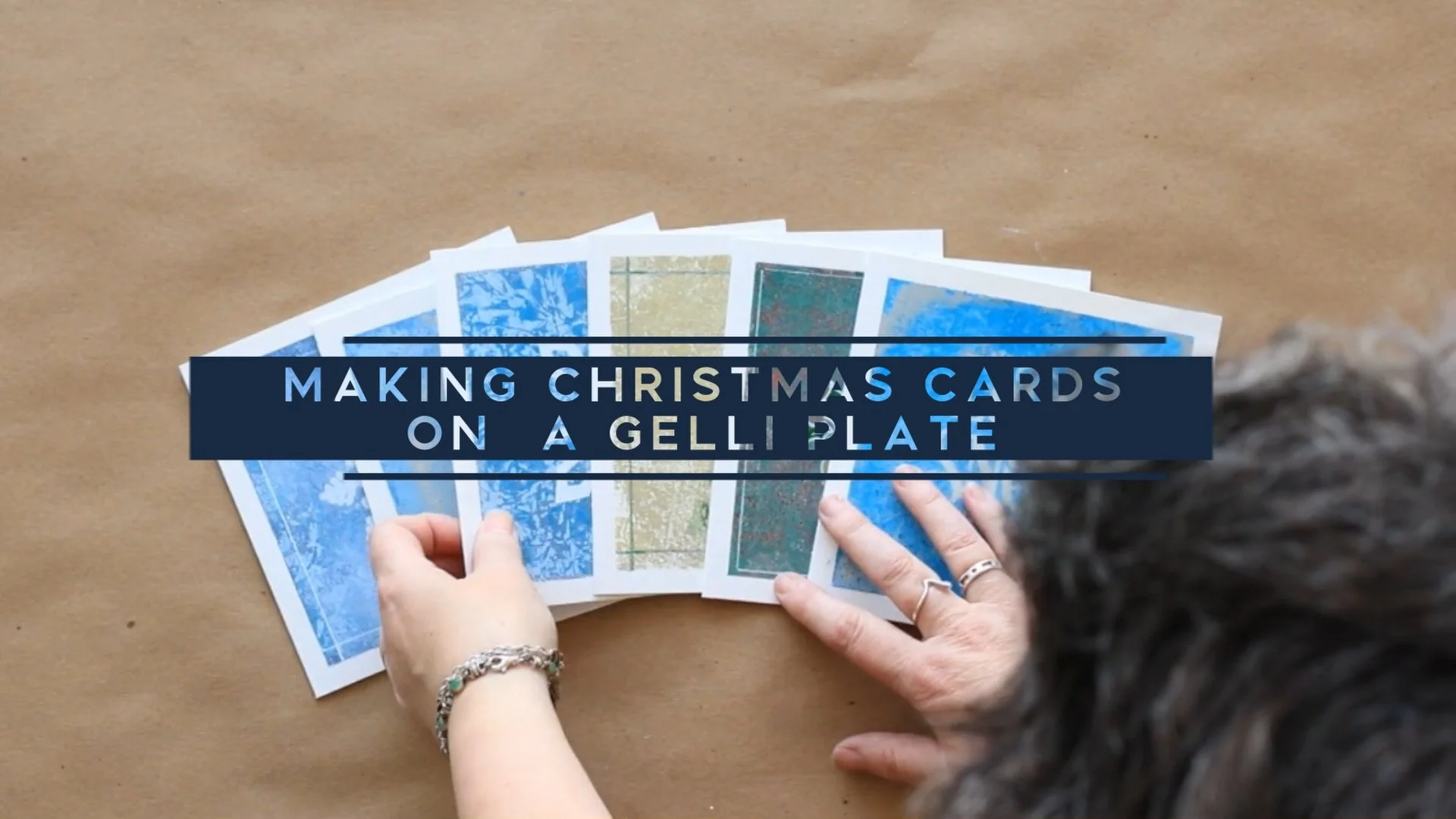 Hands arranging colorful Christmas cards on a wooden surface with a text overlay reading 'Making Christmas Cards on a Gelli Plate.'