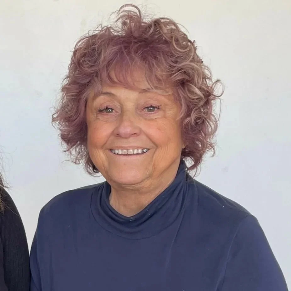 An elderly woman with curly, purple-tinted hair, smiling at the camera, wearing a dark turtleneck top.