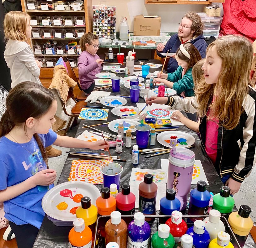 Children and a teacher engaging in a painting activity at a table in an art classroom, with bottles of paint, brushes, and colorful artwork.