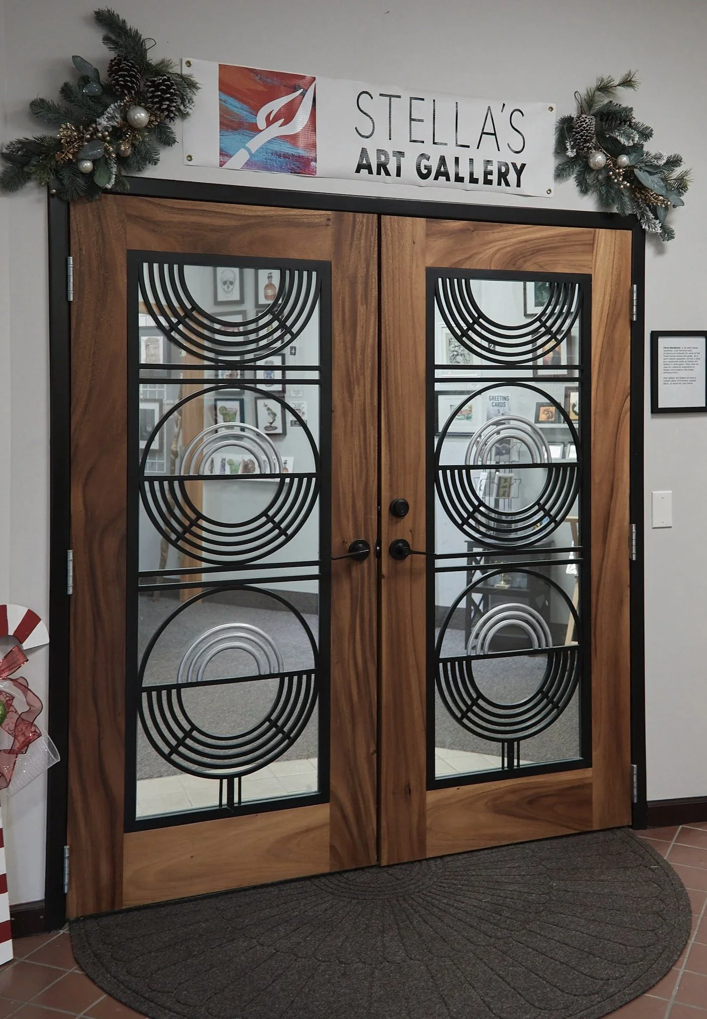 Wooden doors with glass panels featuring geometric black metal designs, flanked by Christmas garlands with ornaments, leading into Stella's Art Gallery.