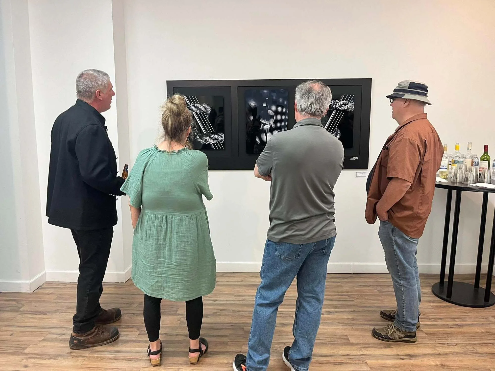 Four people looking at a black and white artwork displayed on a wall in an art gallery. They are standing in a line facing the artwork. To the right, there is a table with bottles and glasses.