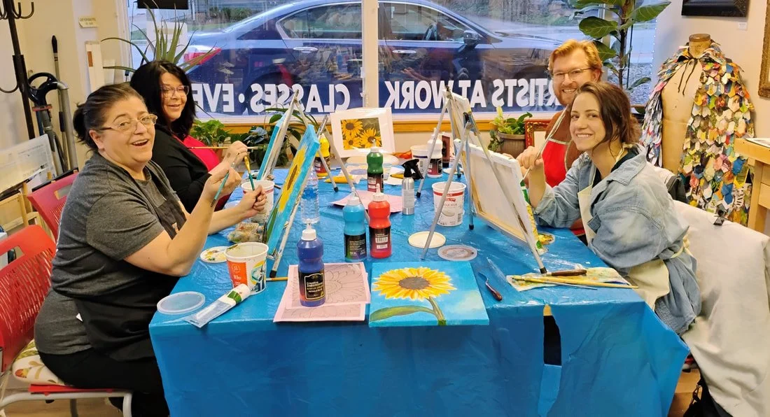 Five people sitting at a painting class, painting sunflowers on canvases, with art supplies on the table, in a bright room.