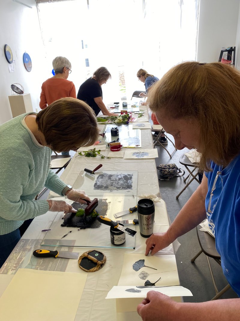 A group of women engaged in printmaking or stamping art activity at a long table in a bright room.