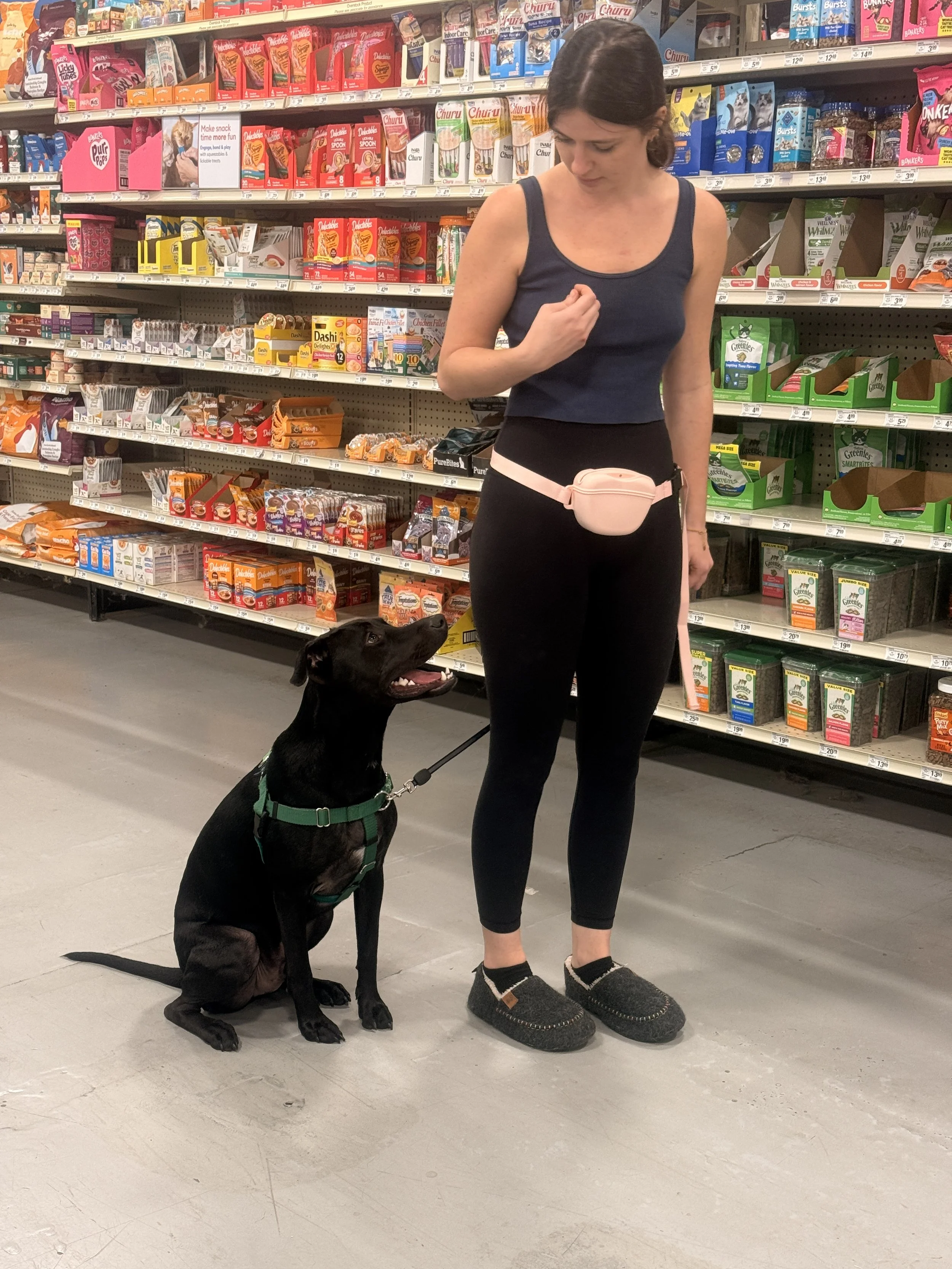 A woman in workout clothes looking down at a black dog sitting beside her in a grocery store aisle, with shelves stocked with snack foods and treats in the background.