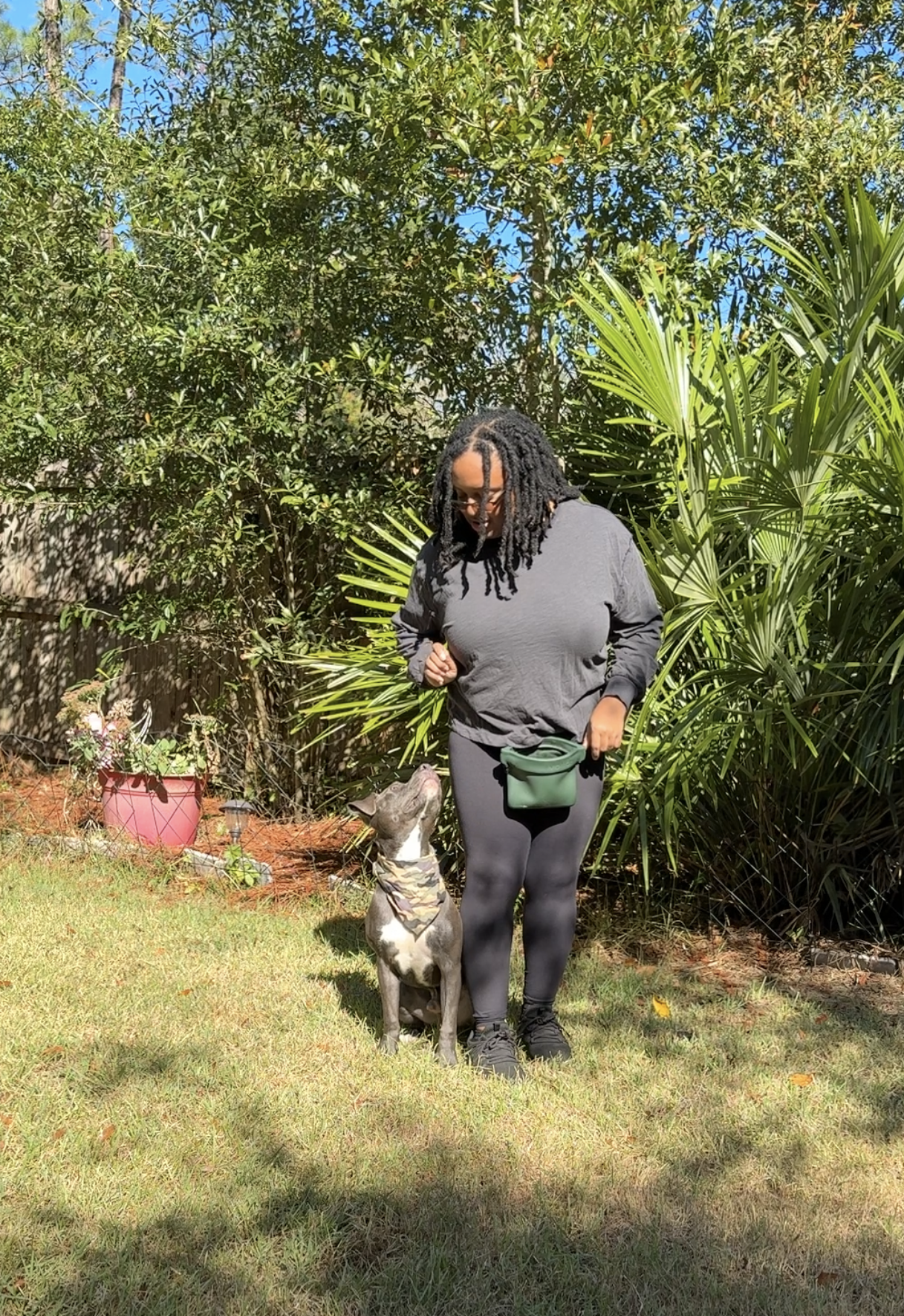 A woman in black athletic clothes standing on grass, looking down at a dog sitting in front of her. The dog is wearing a bandana and looking up at her. There are green plants and bushes in the background, with some pink pots and gardening tools on the ground.
