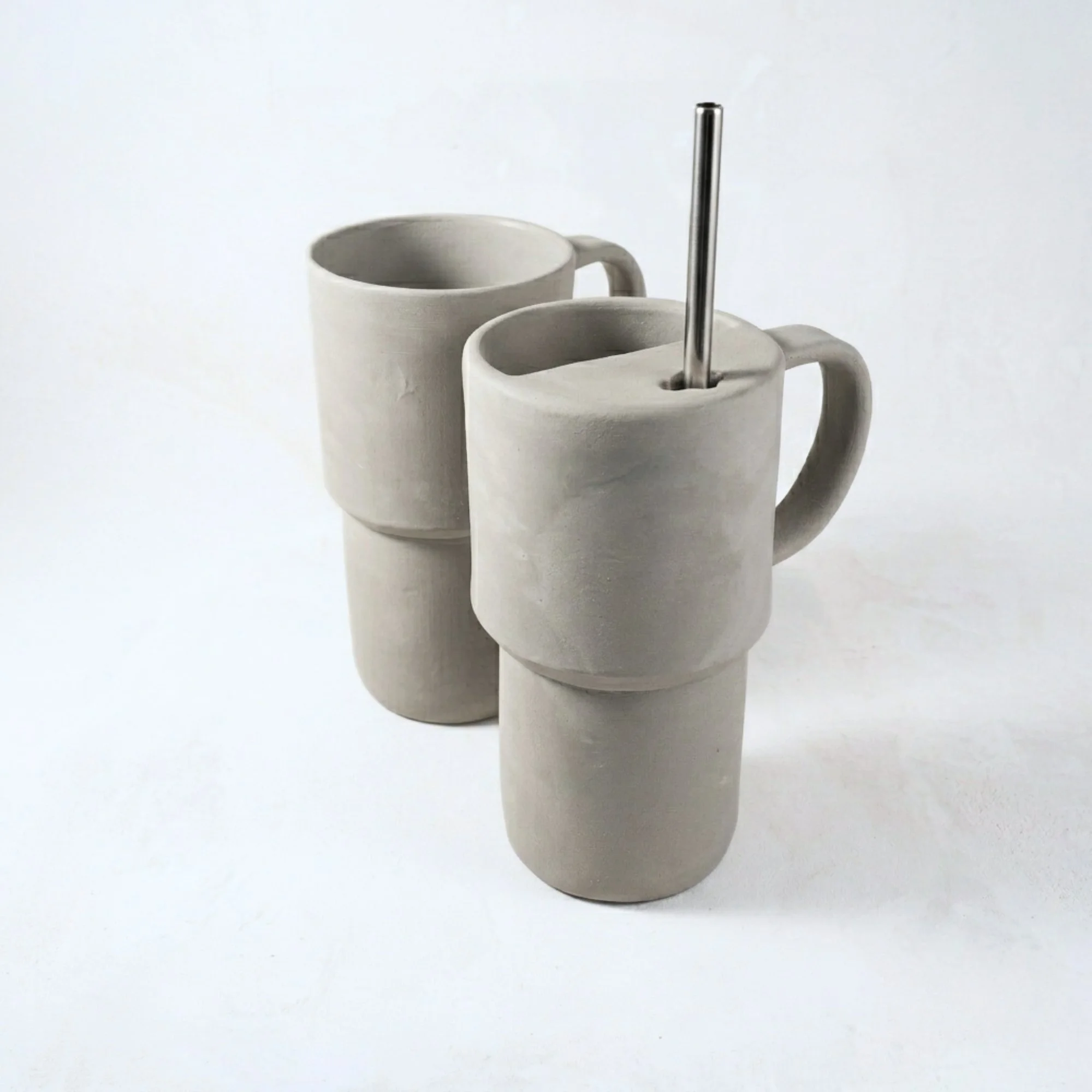 Two light gray ceramic travel mugs with handles. One mug has a stainless steel straw inserted. The mugs have a unique stacked design. The mugs are on a white background. Drinkware.
