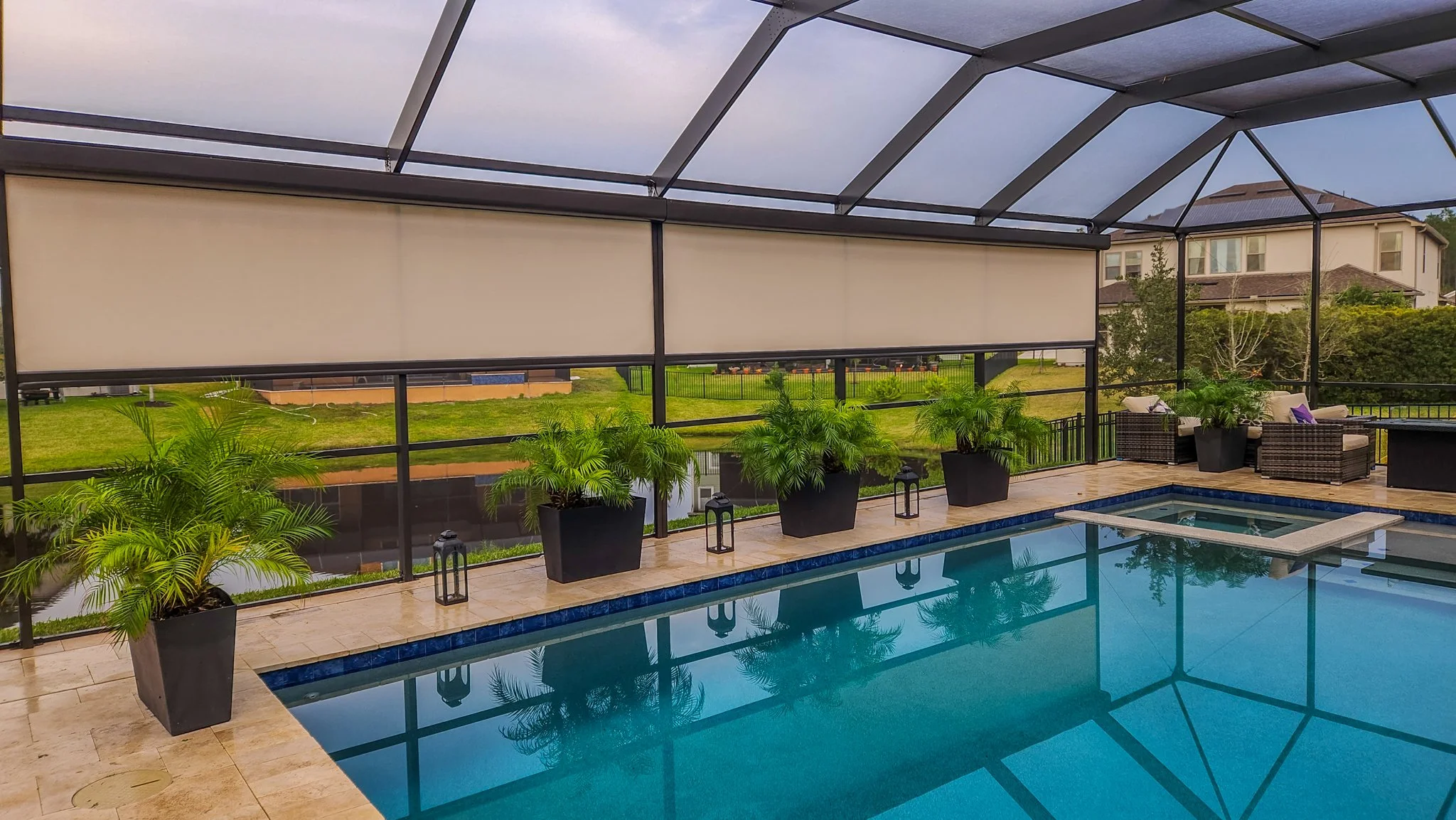 Residential screened-in backyard patio with a swimming pool, potted plants, outdoor furniture, and a view of neighboring houses and greenery.