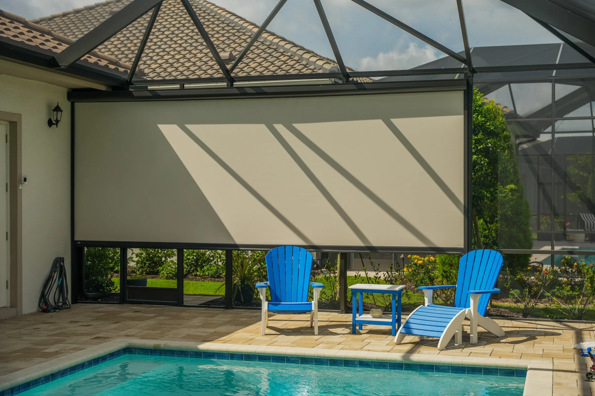 Backyard patio with pool, two blue Adirondack chairs, a side table, and a sunshade with shadows cast on it.