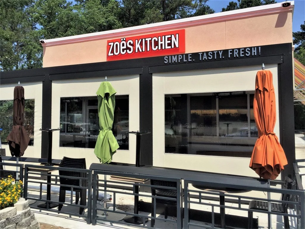 Exterior of Zoës Kitchen restaurant with outdoor seating, colorful umbrellas, and large windows.