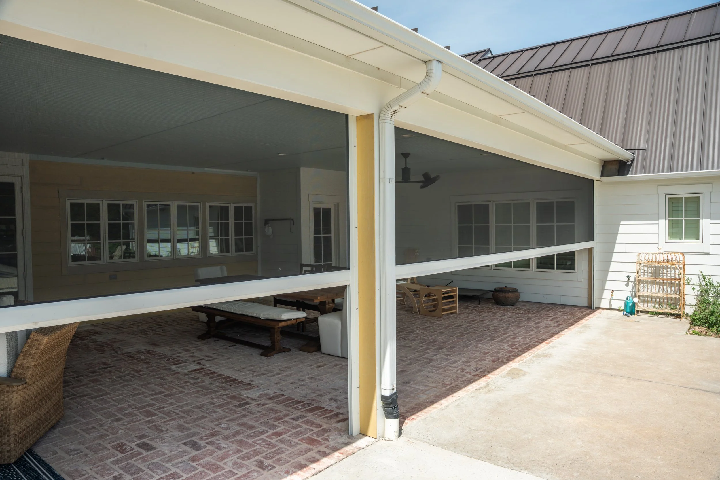 View of a screened-in porch area with brick flooring, outdoor furniture, and windows reflection, attached to a house with white siding.