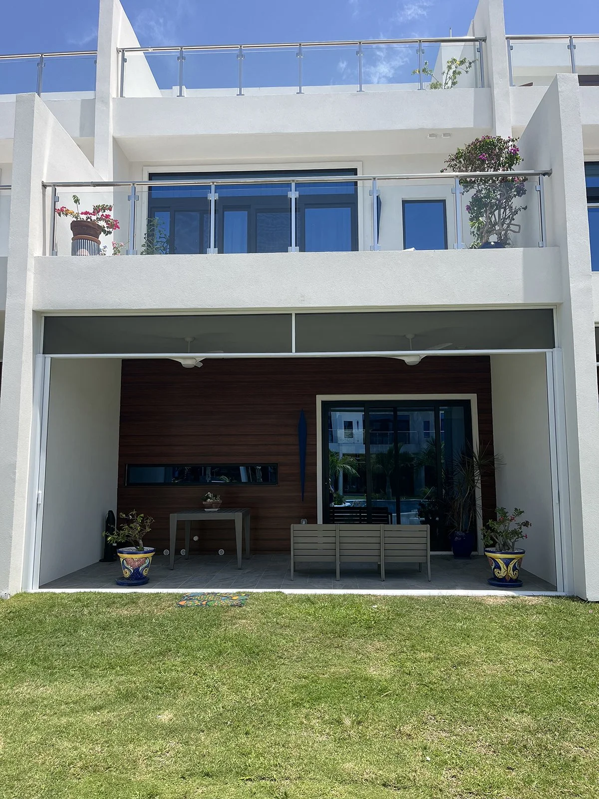 Exterior view of a modern, multi-story apartment or condo building with white walls and glass balconies. The ground floor has a patio with potted plants, a table, and a bench. The upper floors have sliding glass doors and some potted plants on the balconies.