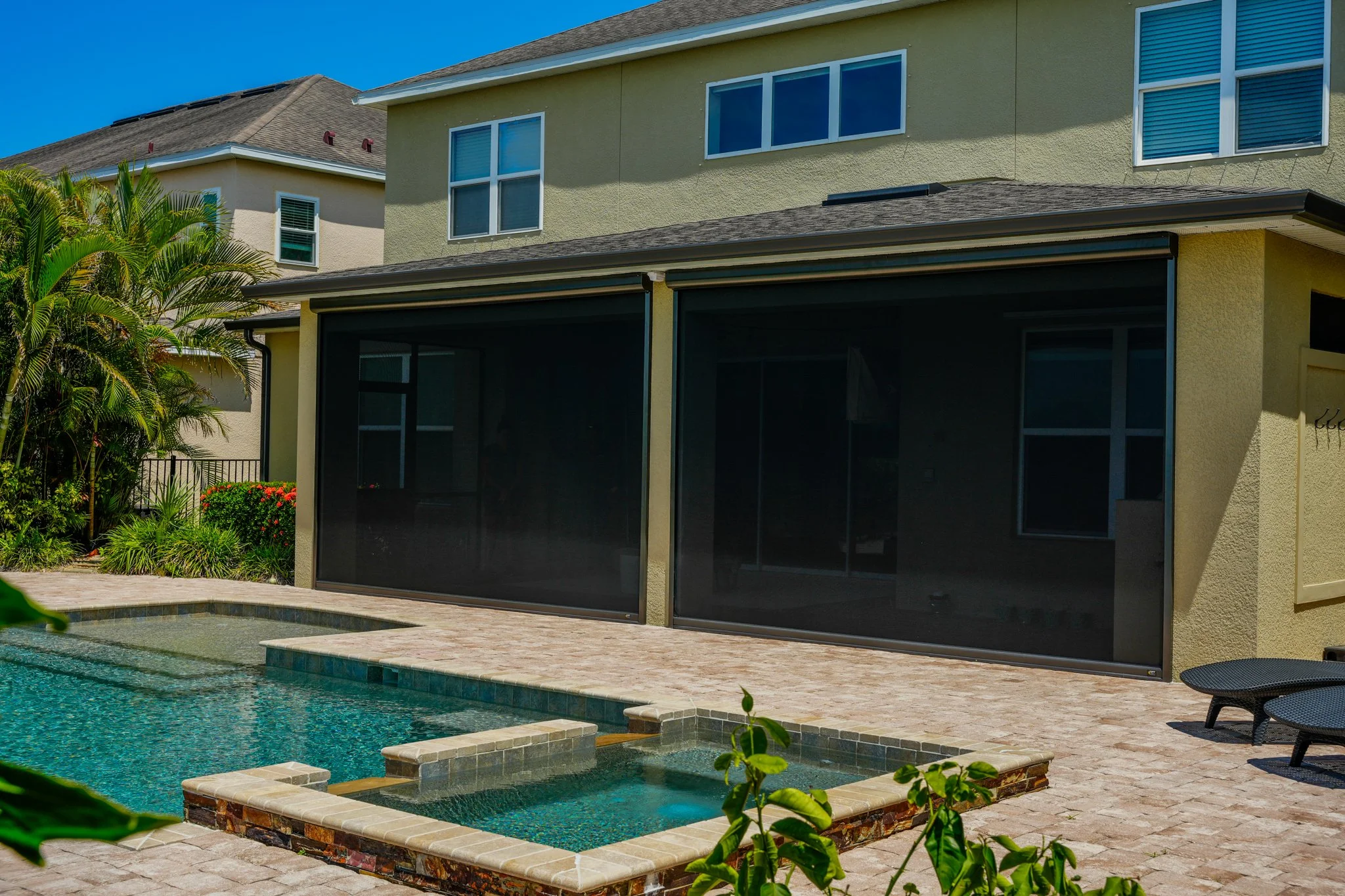 Backyard with swimming pool, hot tub, and screened porch of a yellow house with multiple windows and a neighboring house in the background.