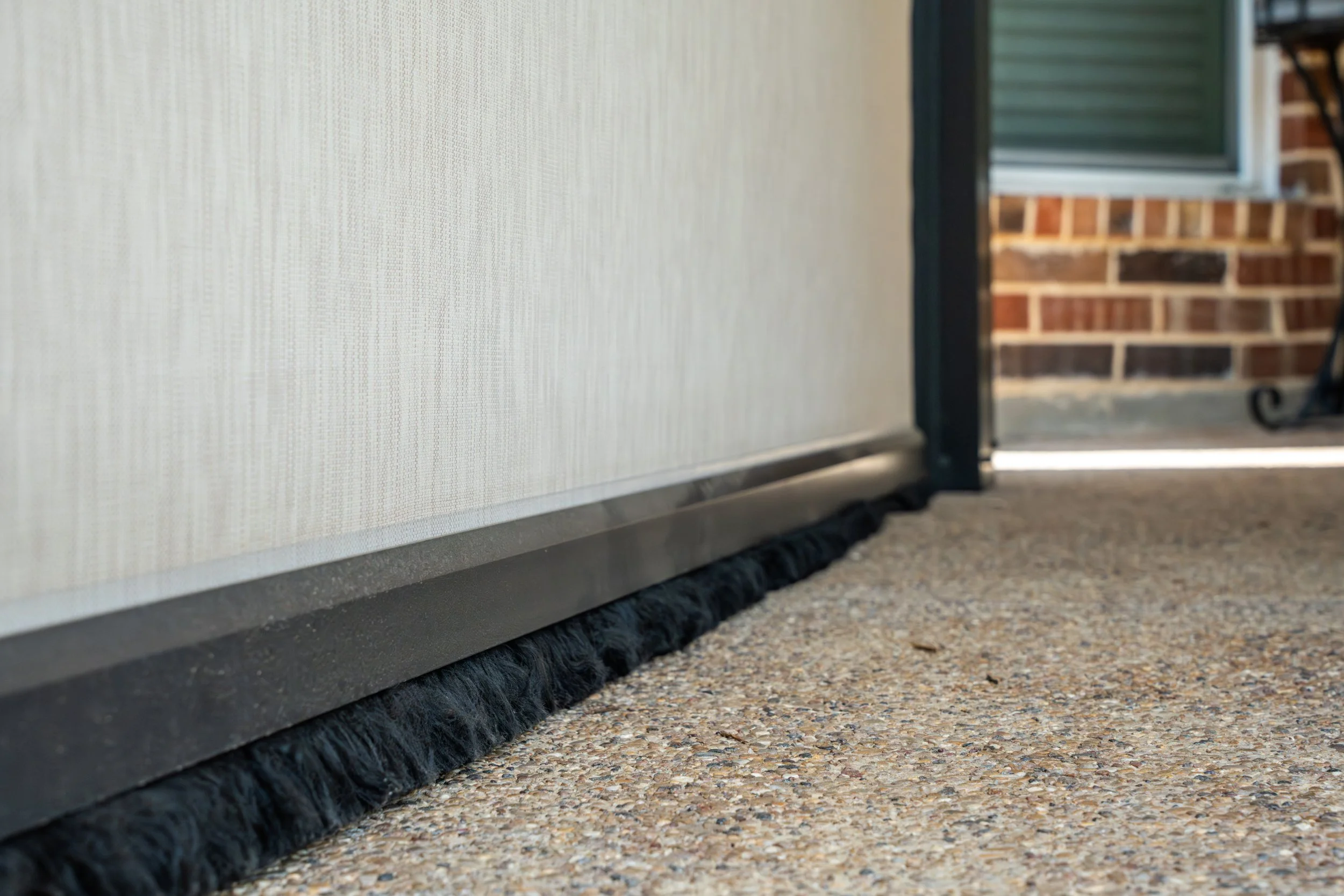 Close-up of a door bottom seal with a black weather stripping on a concrete porch floor, with part of a brick wall and porch in the background.