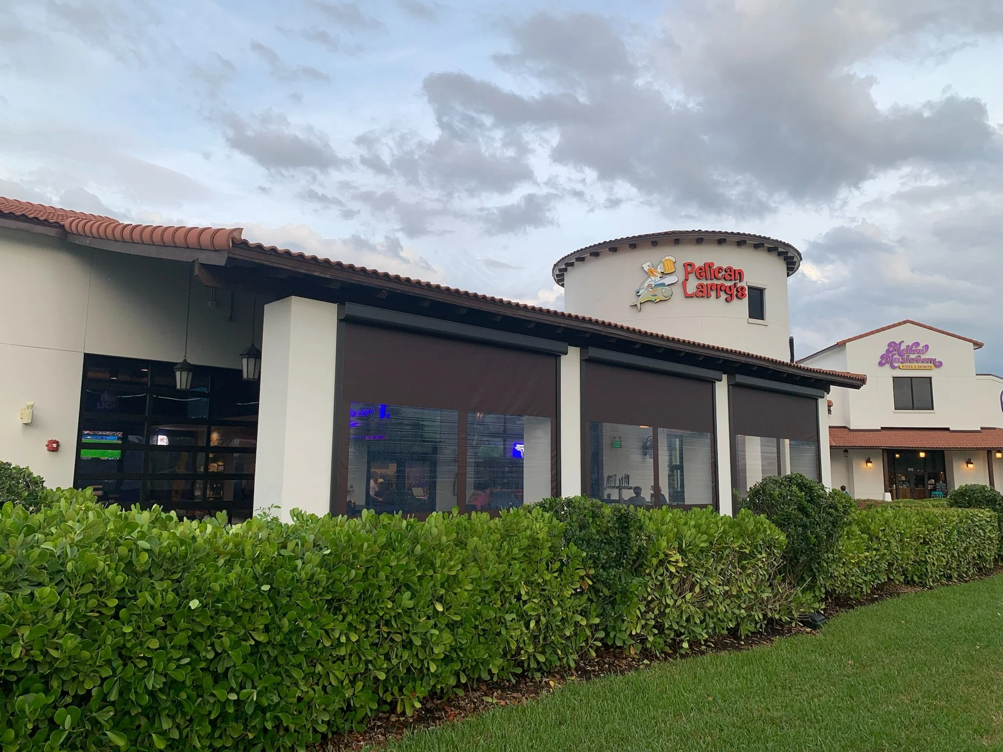 Exterior of Pelican Larry's restaurant with a white building, black window covers, red and purple signs, surrounded by green bushes, under a cloudy sky.