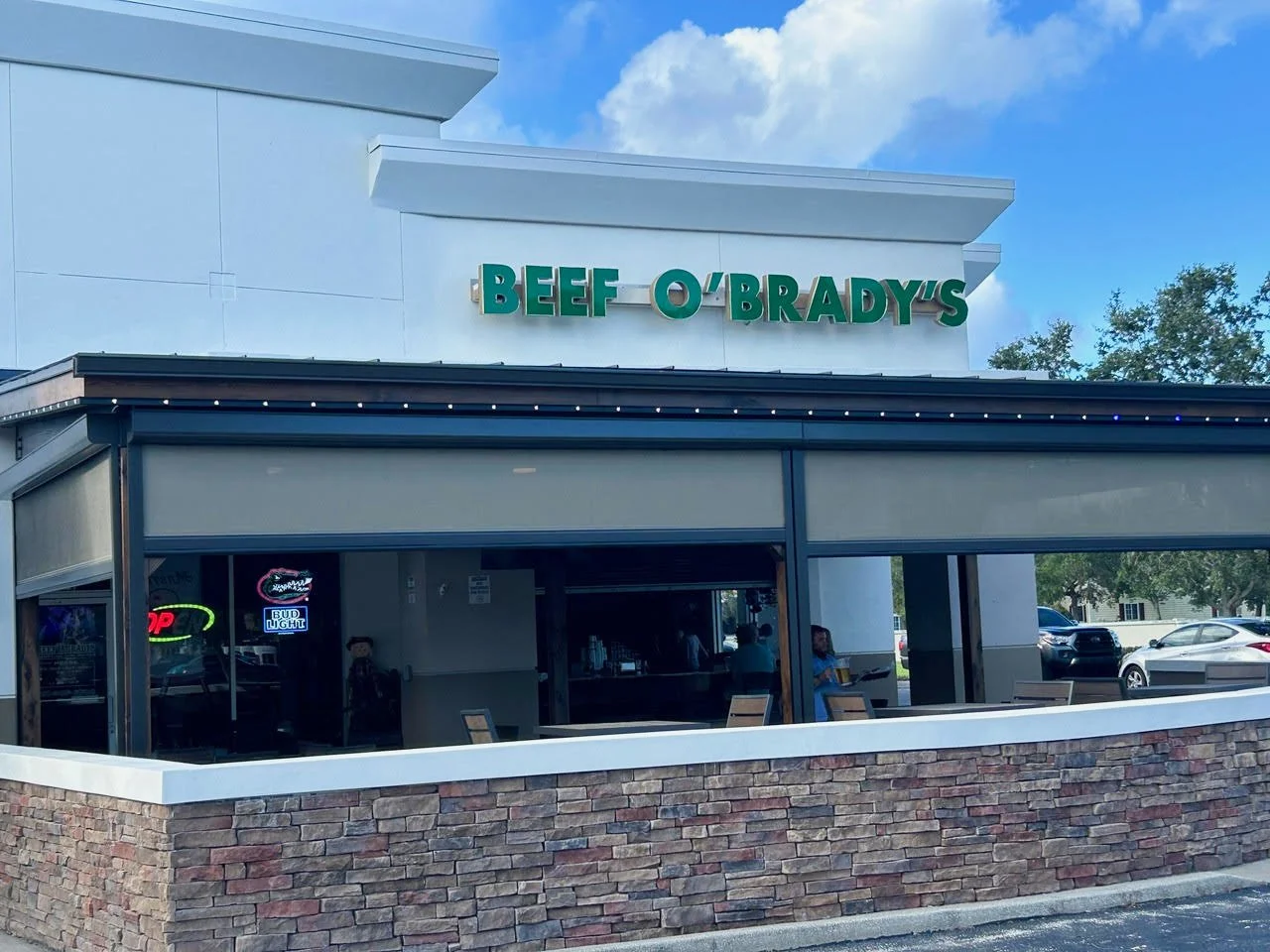 Exterior of Beef O'Brady's restaurant with a sign, small outdoor seating area, and parked cars in the background.