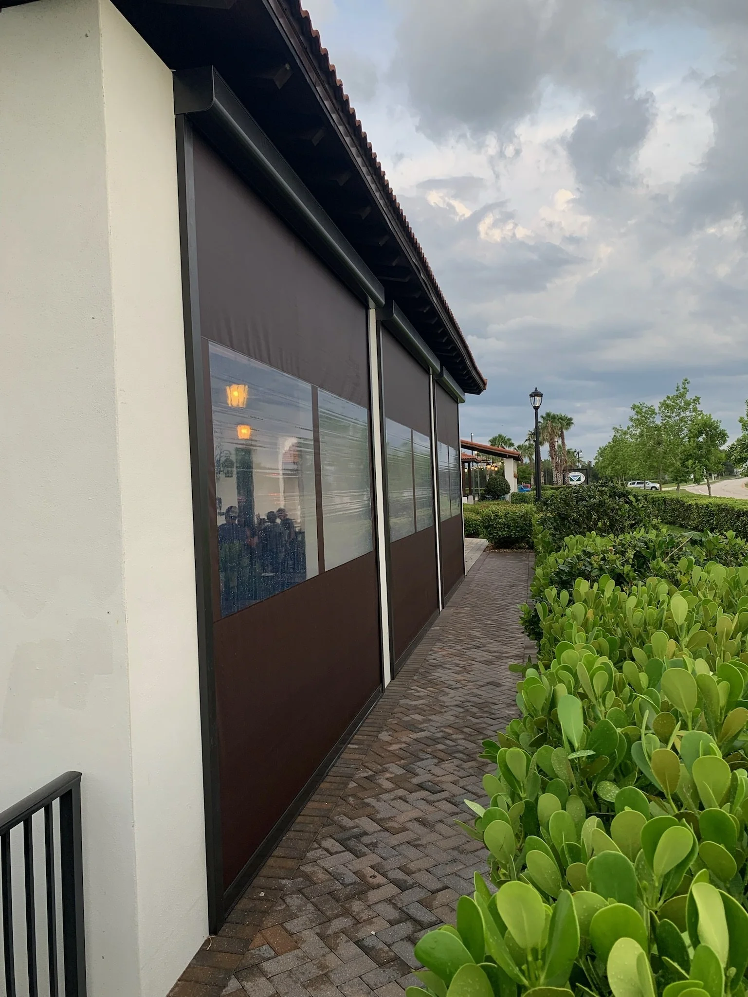 Outdoor sidewalk view with closed dark brown roller shades on the exterior windows of a building, greenery lining the sidewalk, cloudy sky overhead.