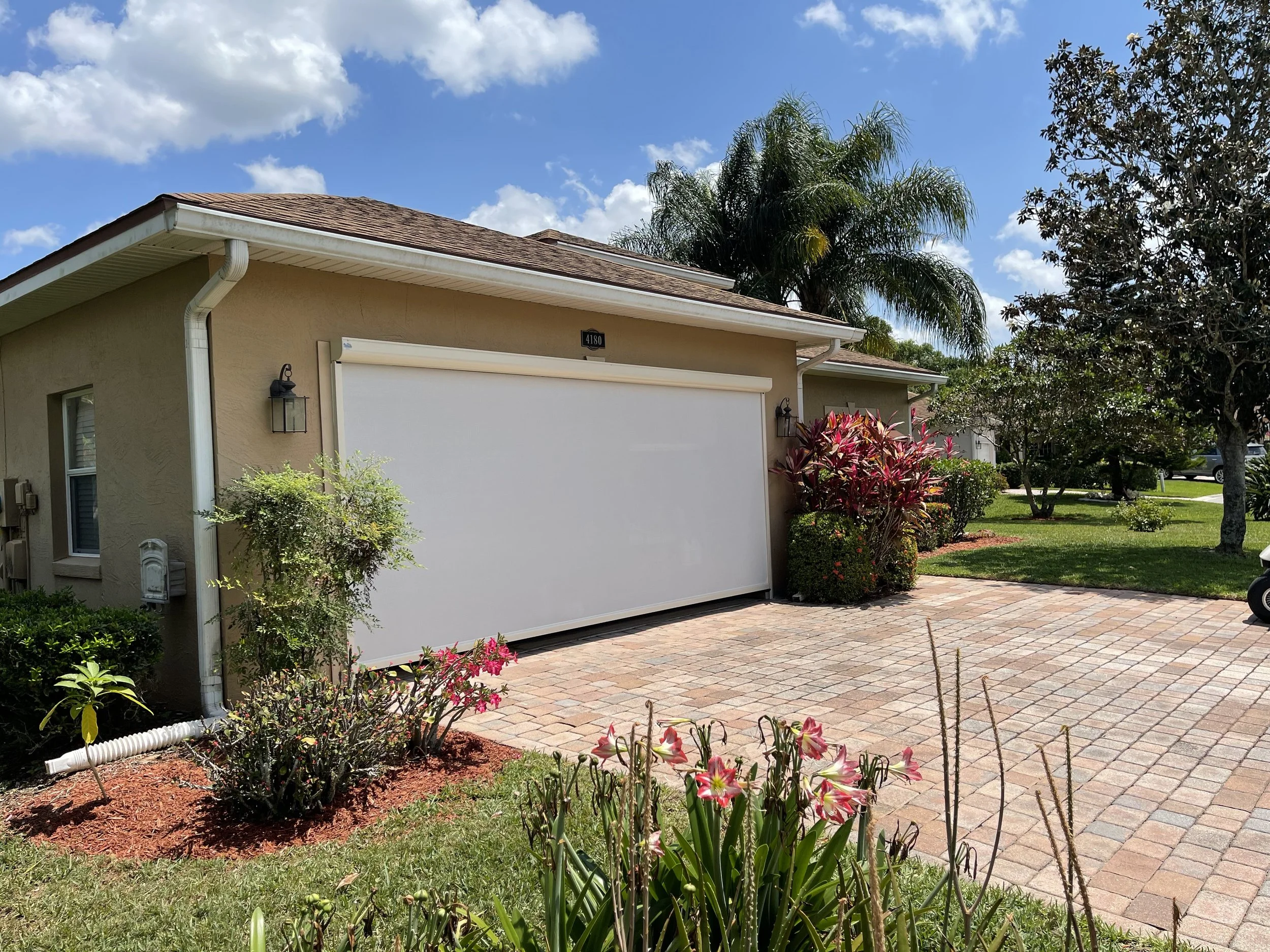 A suburban house with a beige exterior, white garage door, and brick driveway, surrounded by green grass, trees, and flowers, under a partly cloudy blue sky.