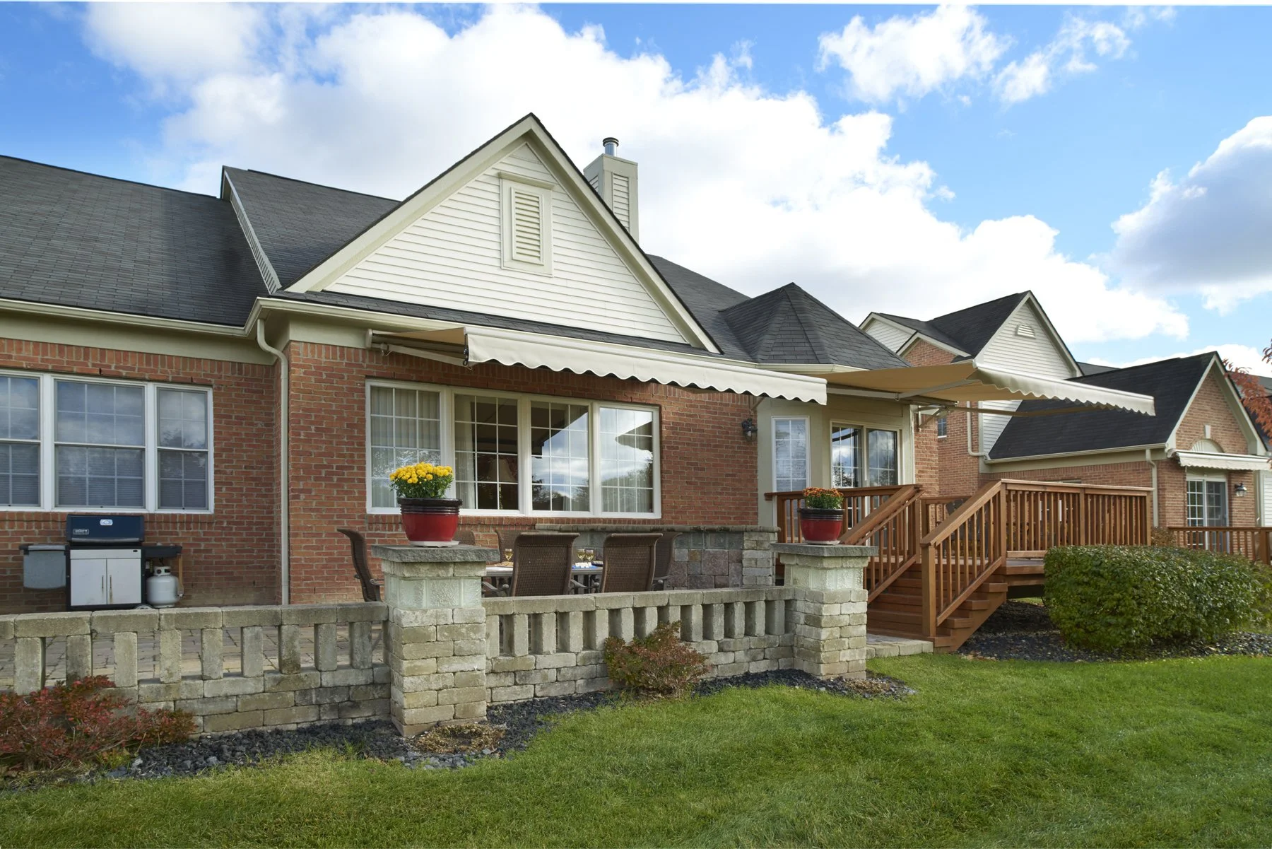 Brick house with a wooden deck, potted flowers, and outdoor furniture under a blue sky.