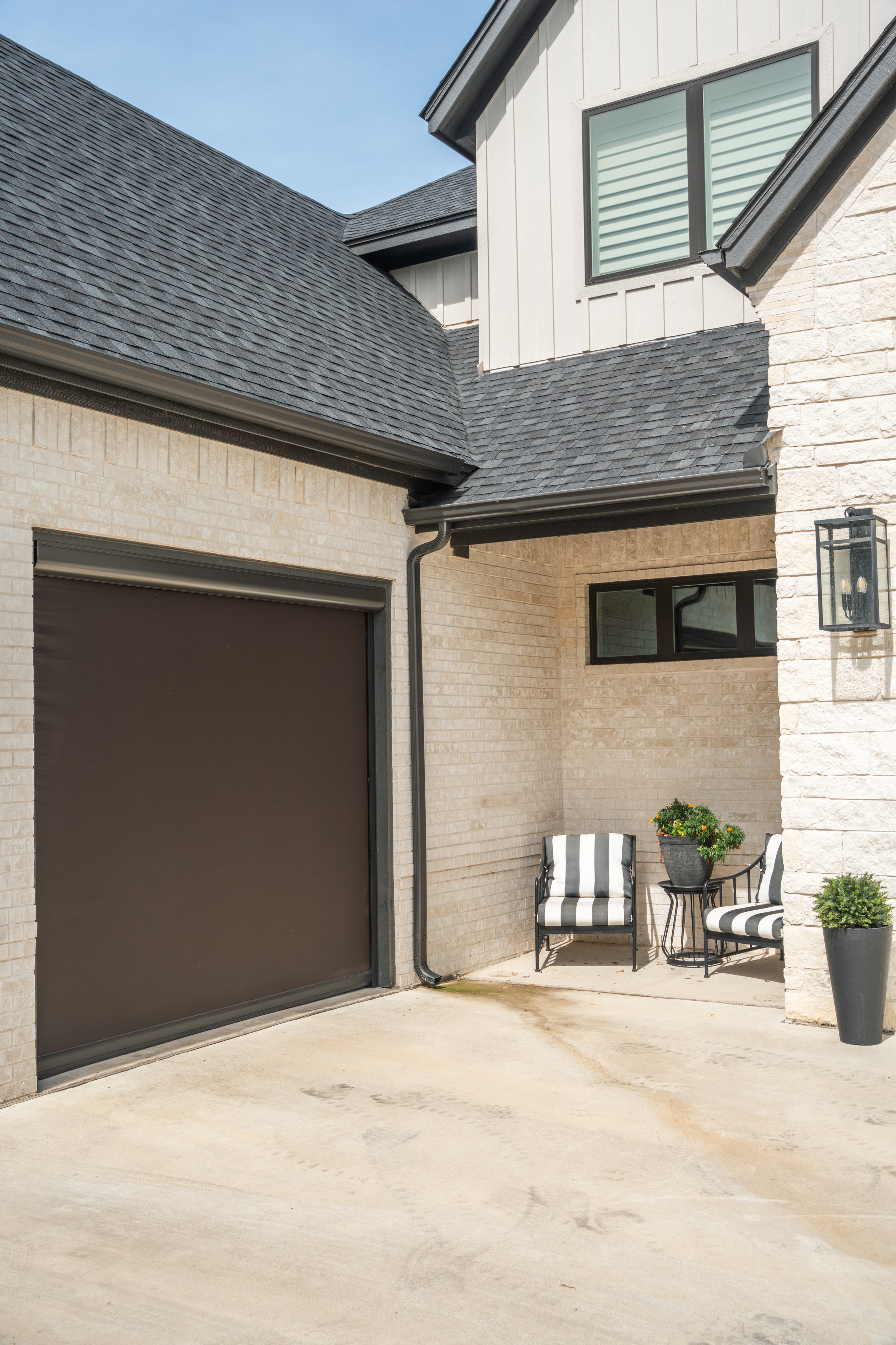 A modern house exterior with a beige brick wall, black window, black light fixture, black chairs with striped cushions, and potted plants