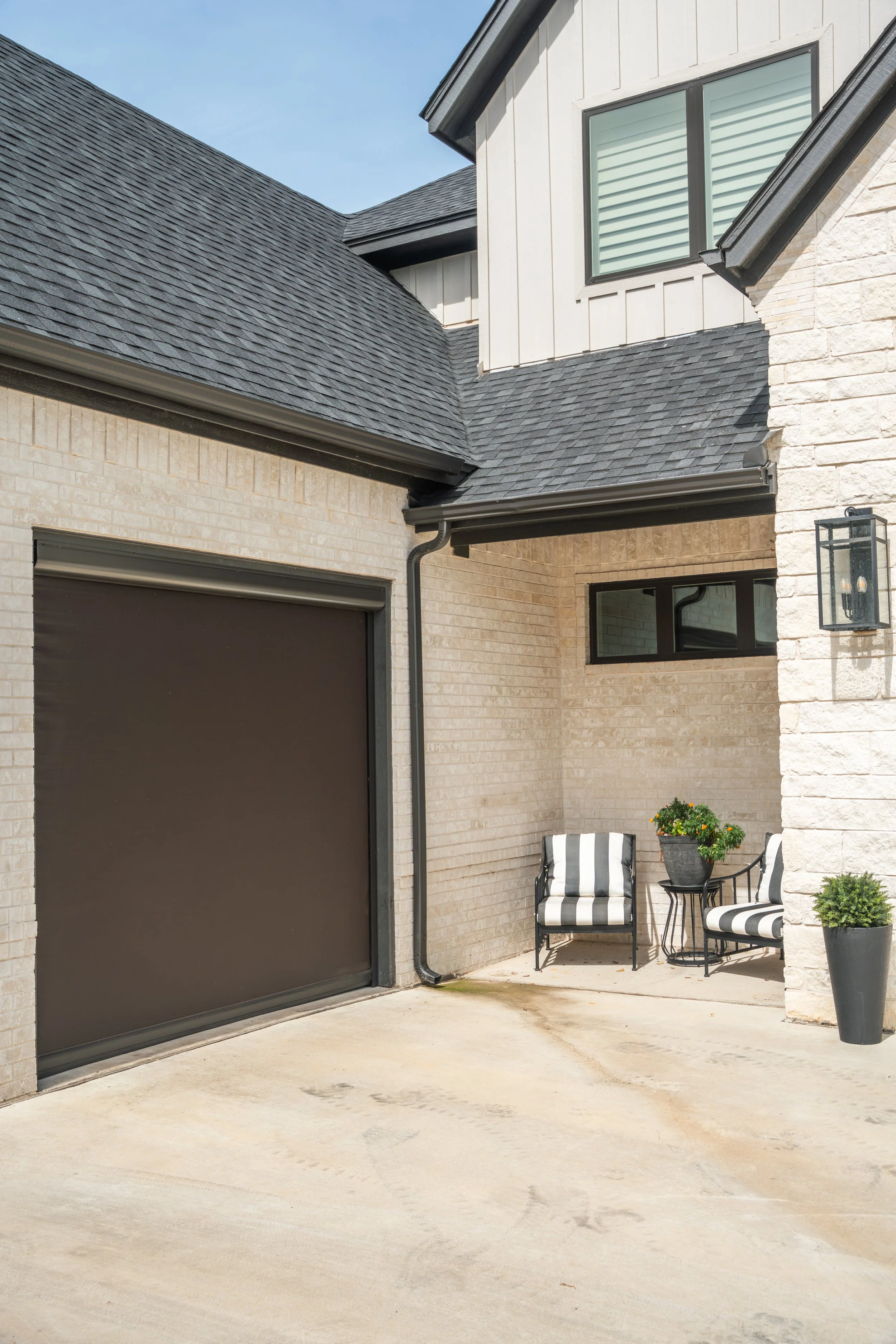 Residential house exterior with garage door, patio seating area with two chairs, a small table, potted plants, window, and wall-mounted light fixture.