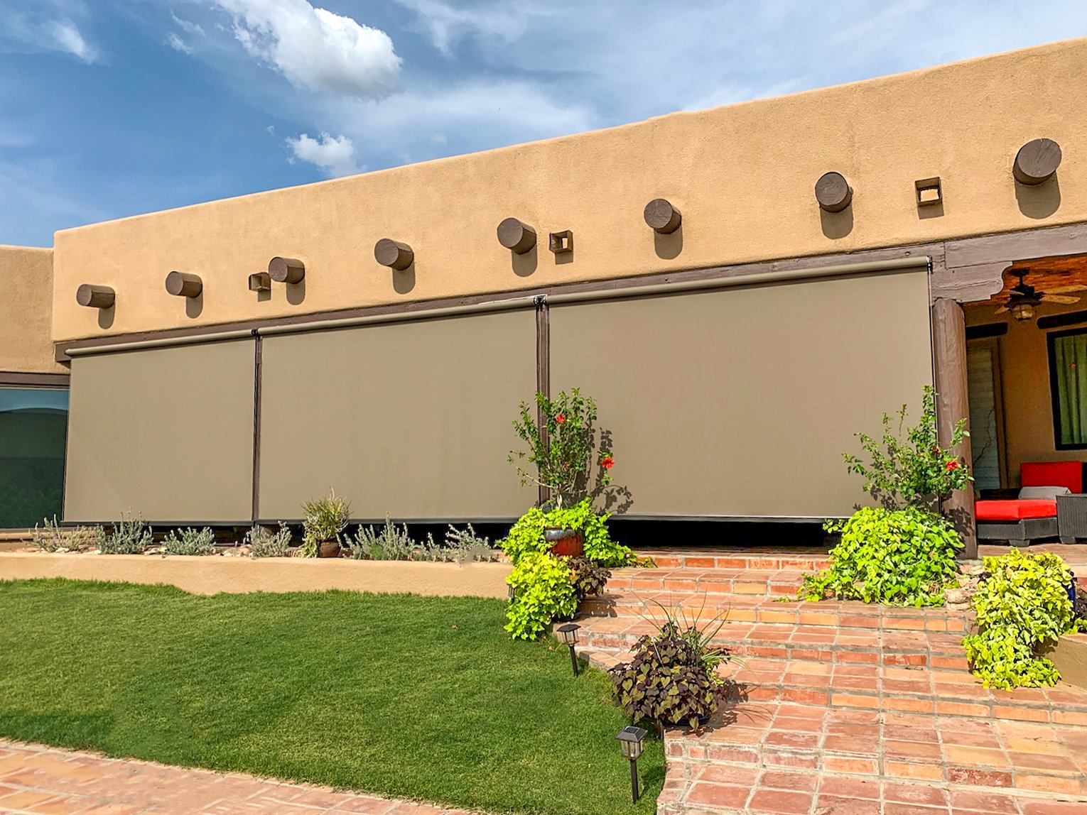 Patio with brick steps, green lawn, and plants in front of a beige building with beige retractable awnings.