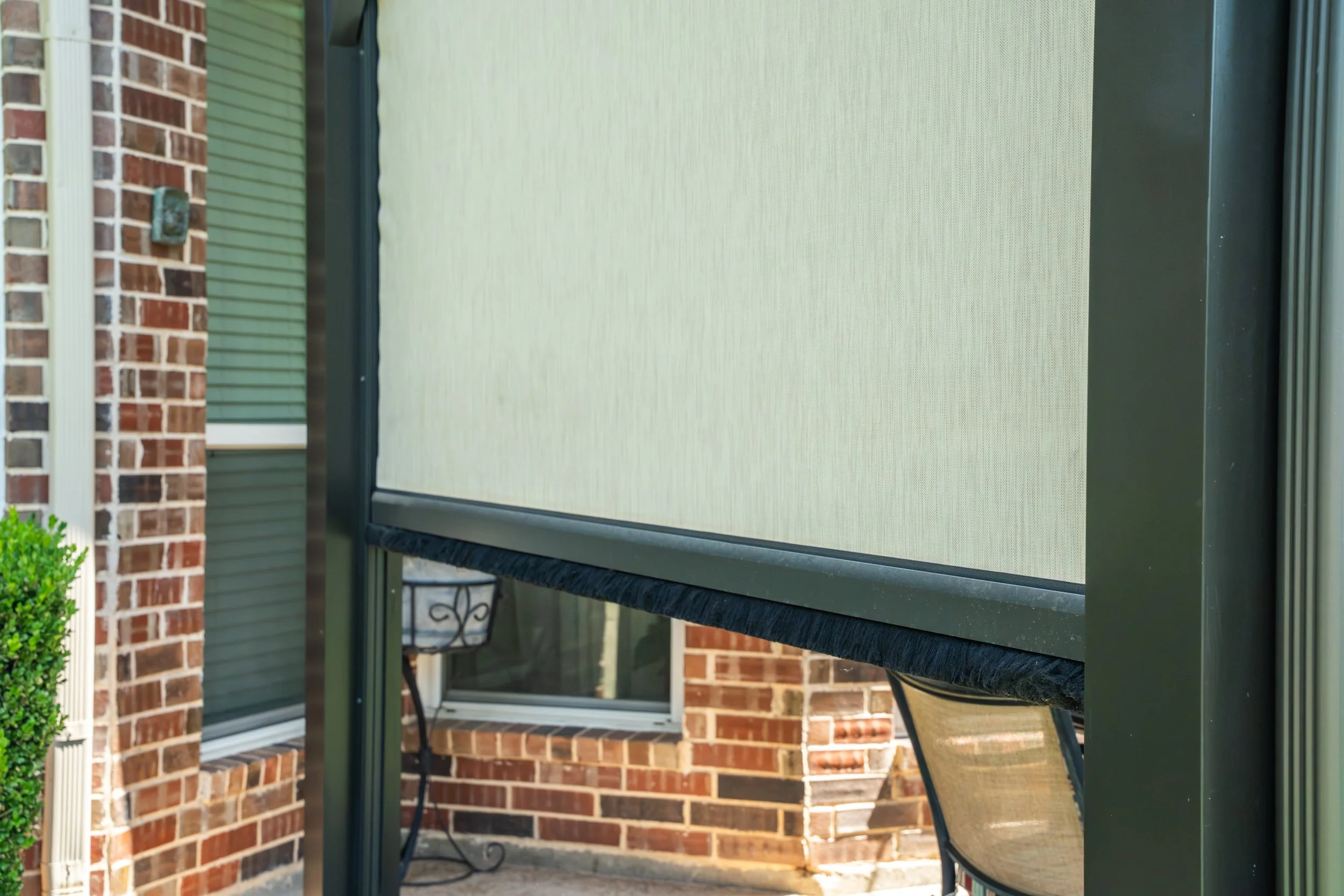 A beige outdoor retractable screen or shade on a patio, with a brick house and greenery in the background.