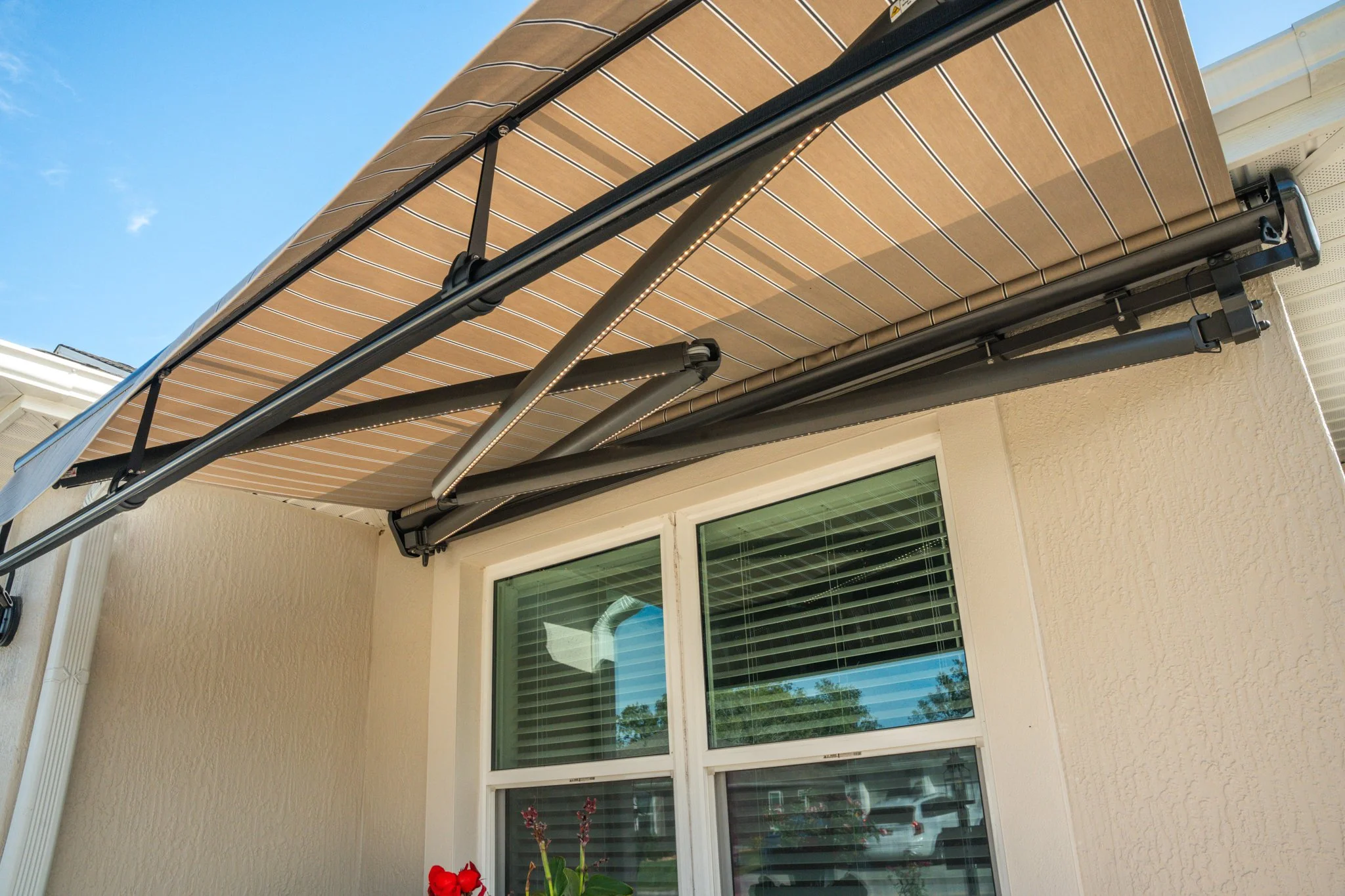 Retractable beige striped awning extended above a window on the exterior of a house with beige textured walls and a clear blue sky.