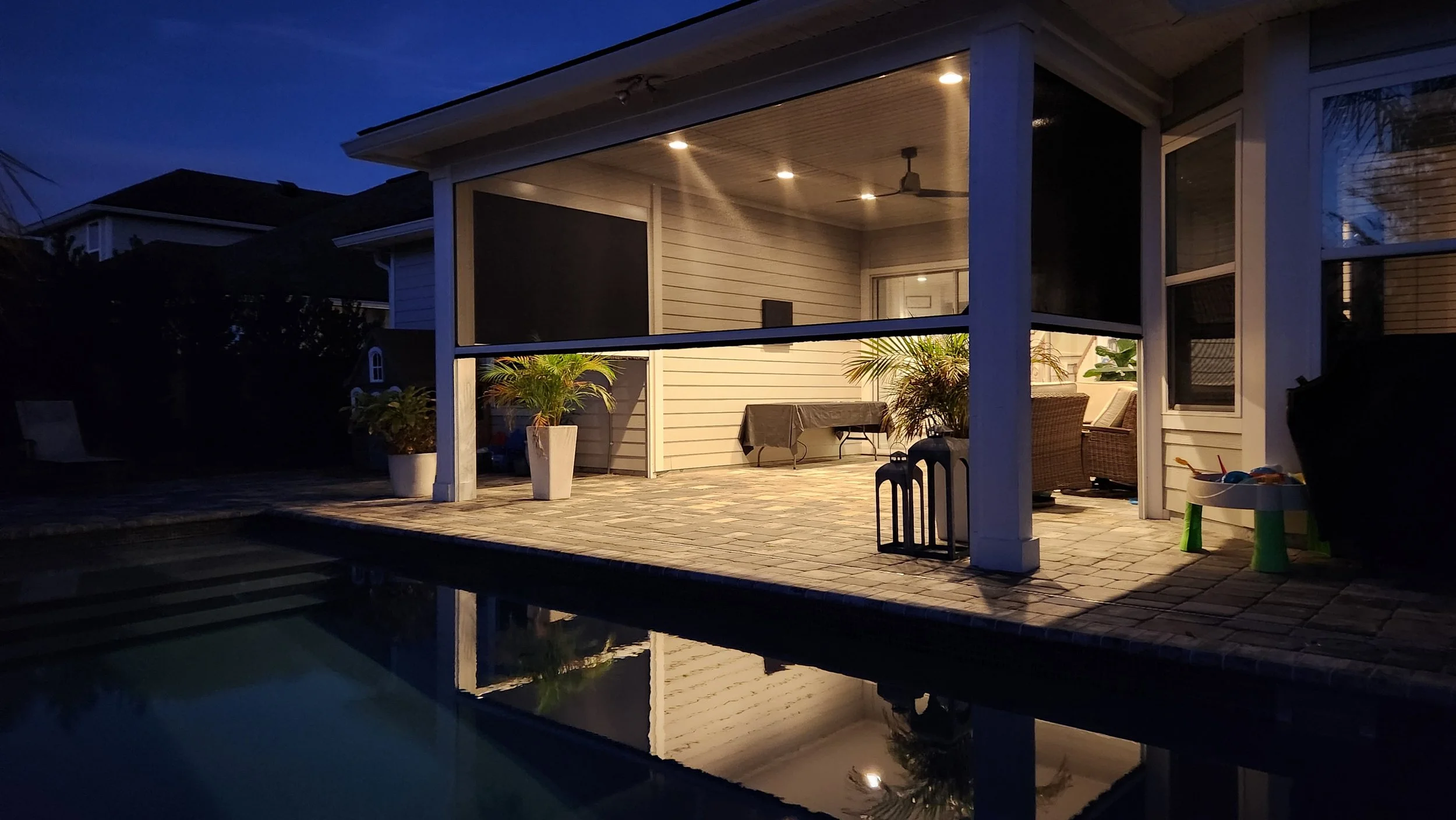 A covered backyard patio area at dusk with outdoor furniture, potted plants, and decorative lanterns, reflected in a swimming pool.