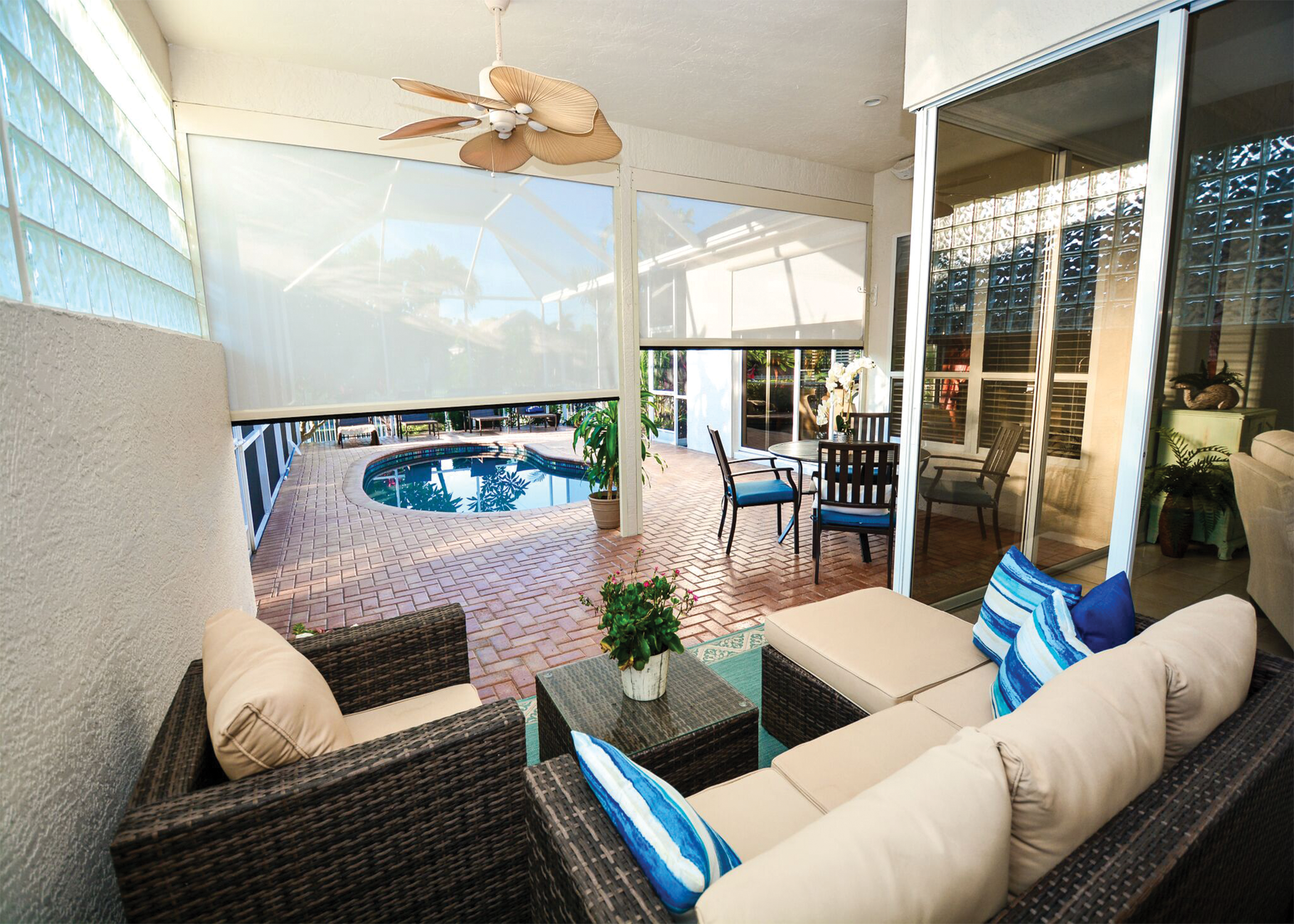 Enclosed patio with wicker sofa and blue striped pillows, small table with plant, sliding glass door views of outdoor pool area with chairs and umbrellas, ceiling fan with tropical leaf design.