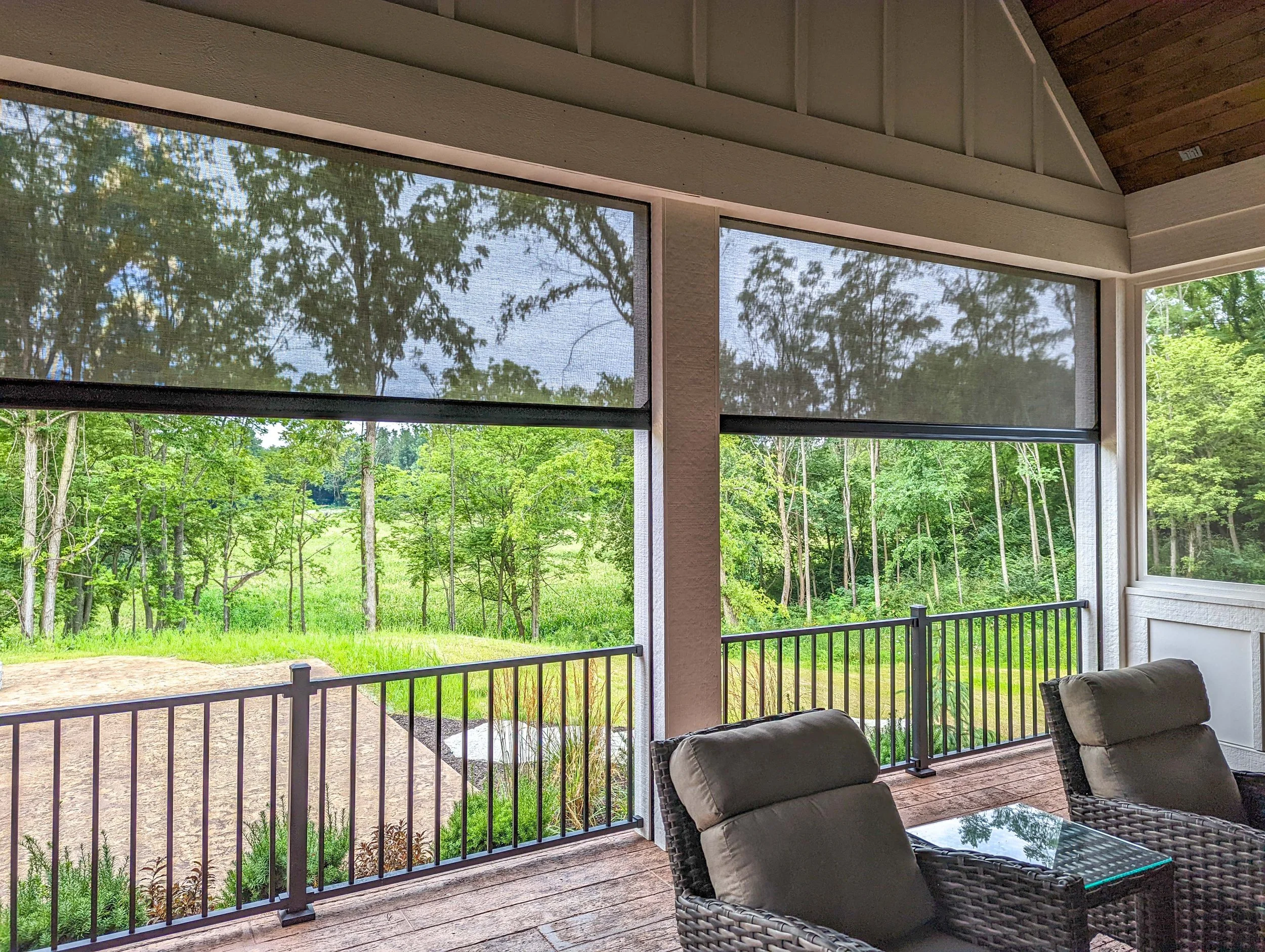 View from screened porch showing green trees, grass, and a small pond or stream in the background, with patio furniture and railing in the foreground.