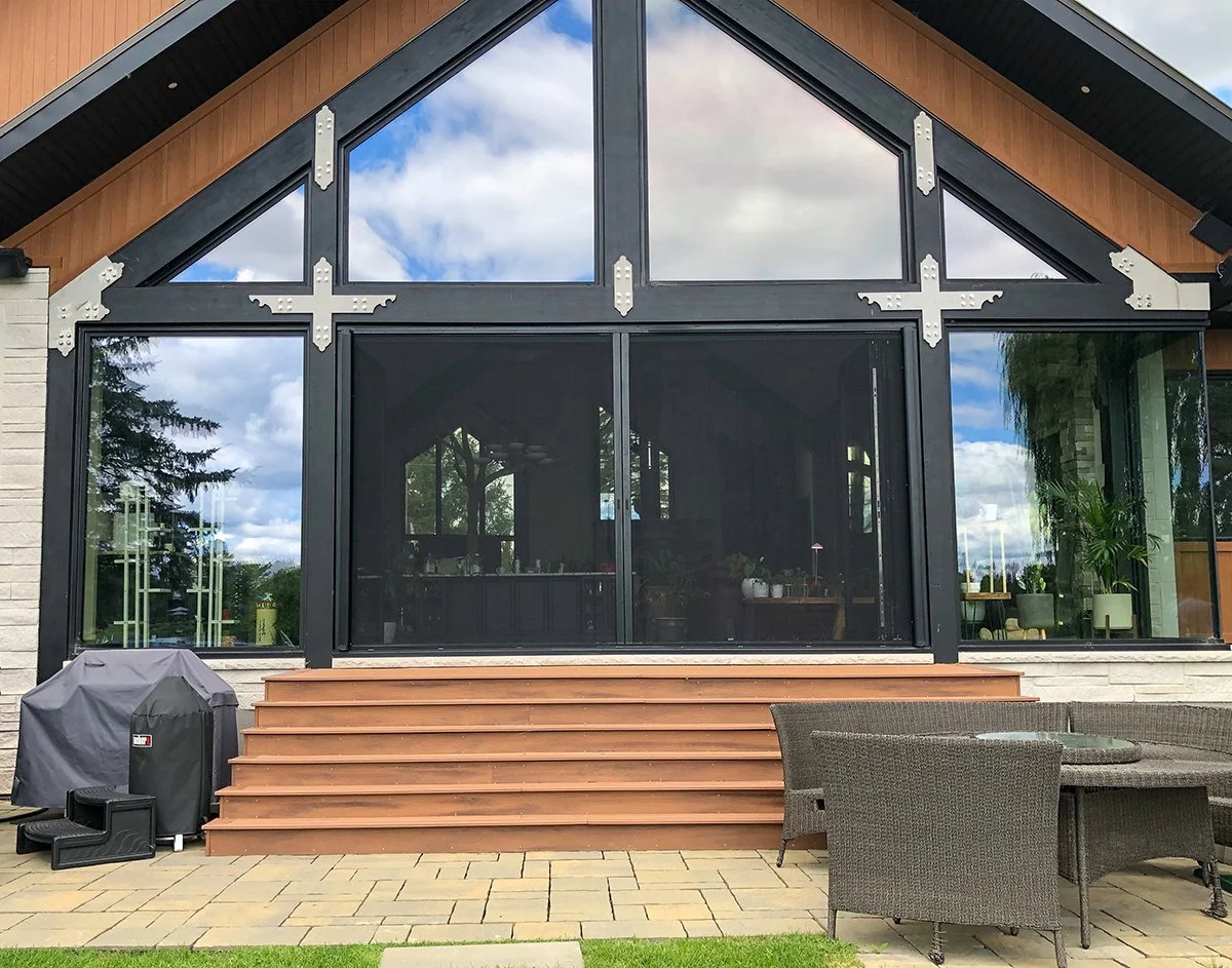 Modern house with large glass windows and doors, wooden steps leading up to the entrance, outdoor patio with wicker furniture, and a grill covered with a protective cover.