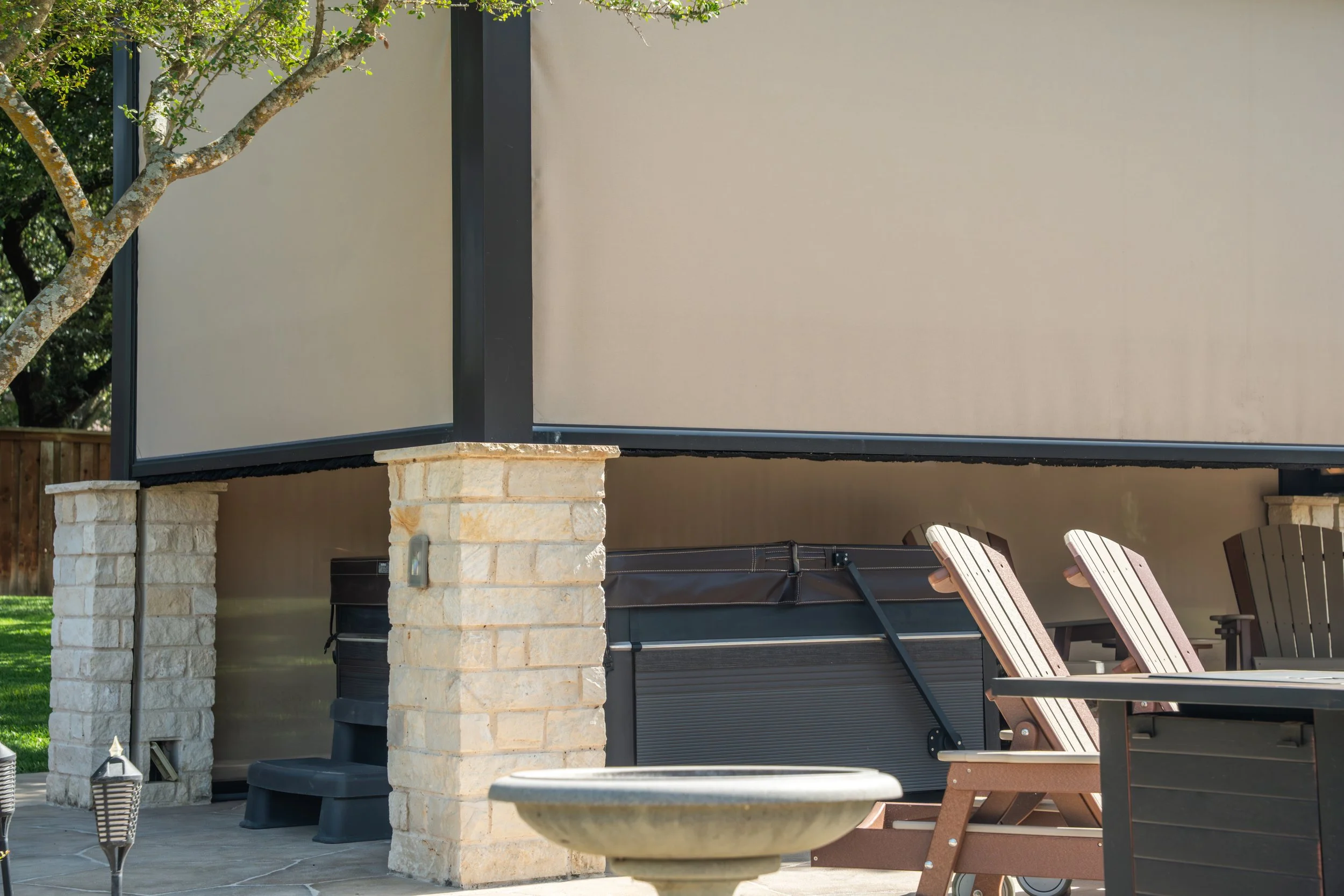A patio area with stone pillars, Adirondack chairs, a hot tub, and outdoor lighting fixtures, with a house and trees in the background.