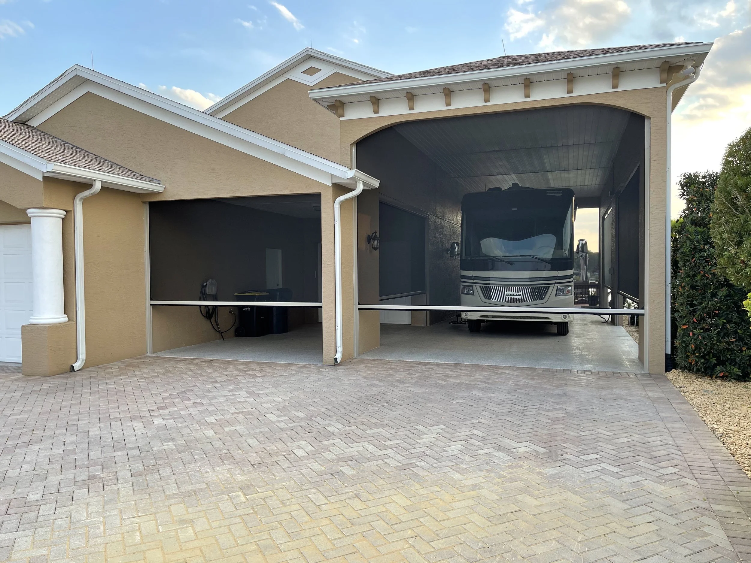 A house with an attached garage that has a screened-in carport. Inside the carport, there is a recreational vehicle (RV) parked. The garage has a driveway and a small area with scant landscaping on the right.