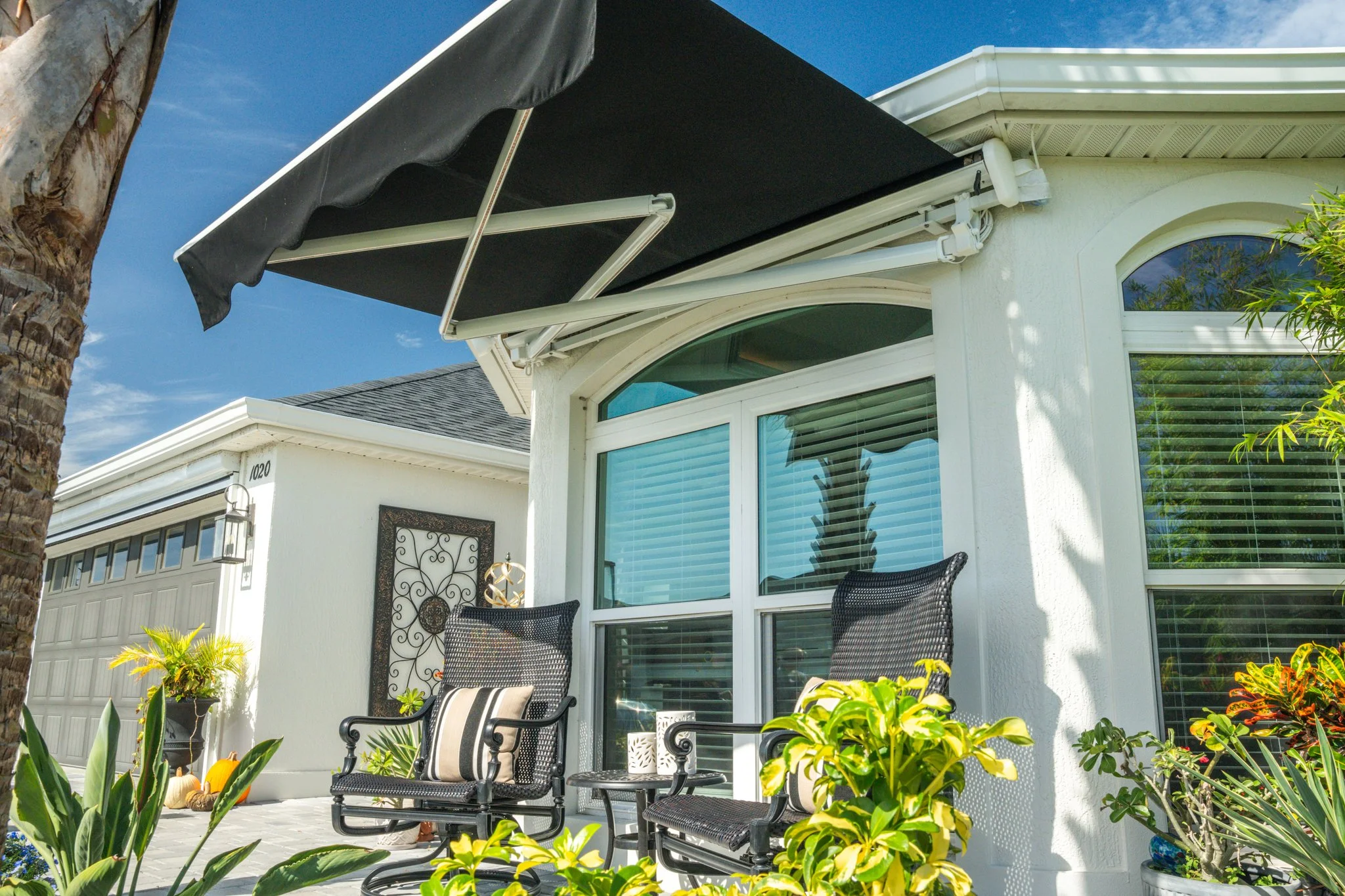 A white house with large windows, a black awning, two black chairs with striped pillows on a patio, surrounded by green plants and a palm tree, under a blue sky.