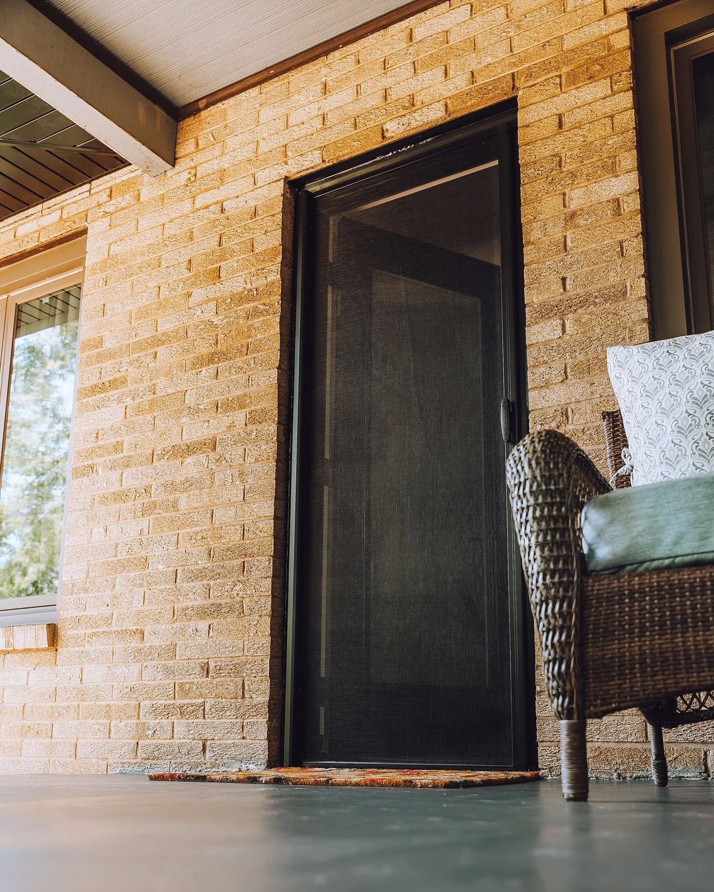 Close-up of a black screen door with brick exterior wall, area rug at door, wicker chair with cushion and pillow, sunlit window, and ceiling with wood paneling.