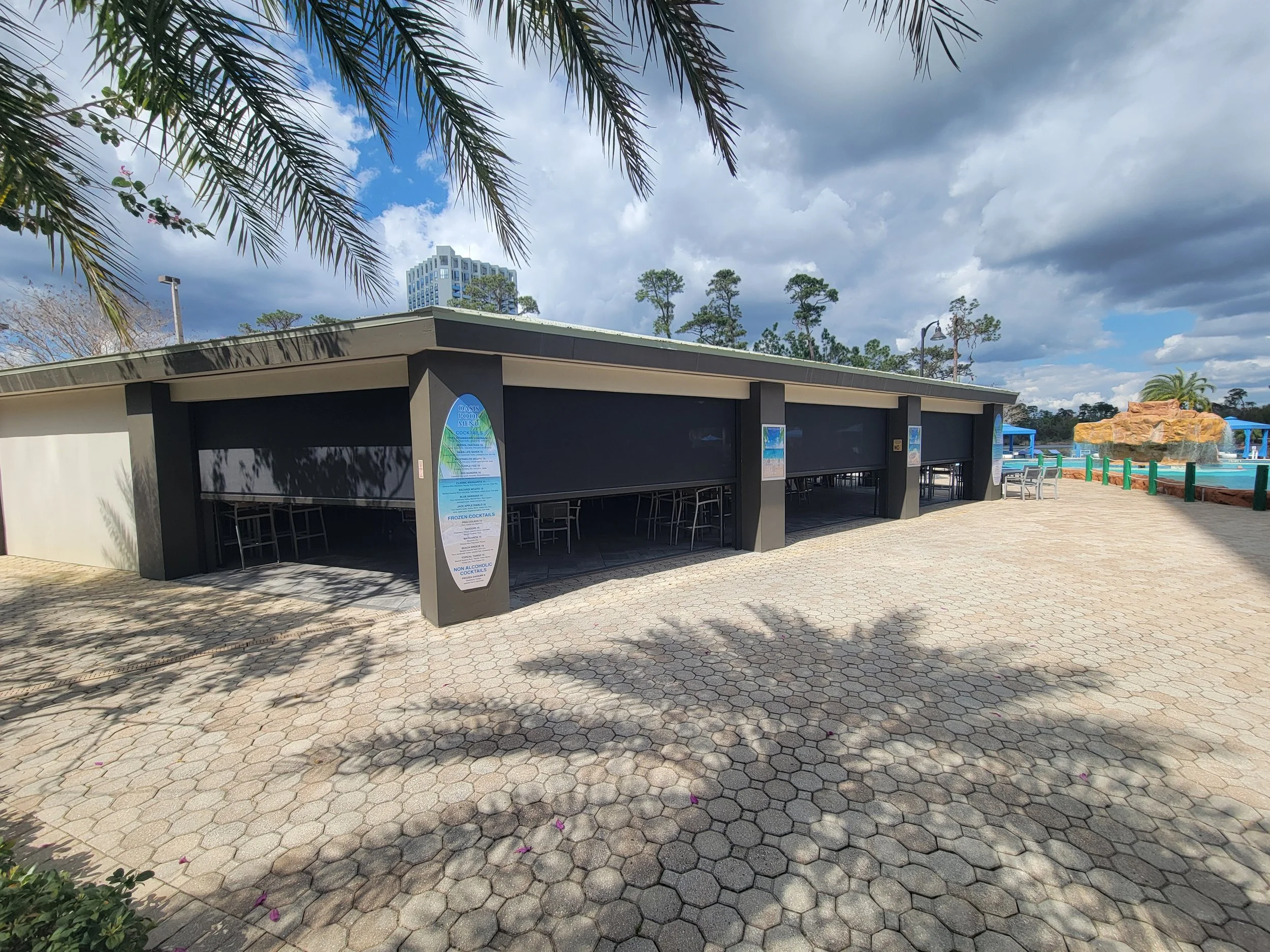 Empty outdoor poolside bar with closed shutters, surrounded by hexagonal paving stones, trees, and cloudy sky.