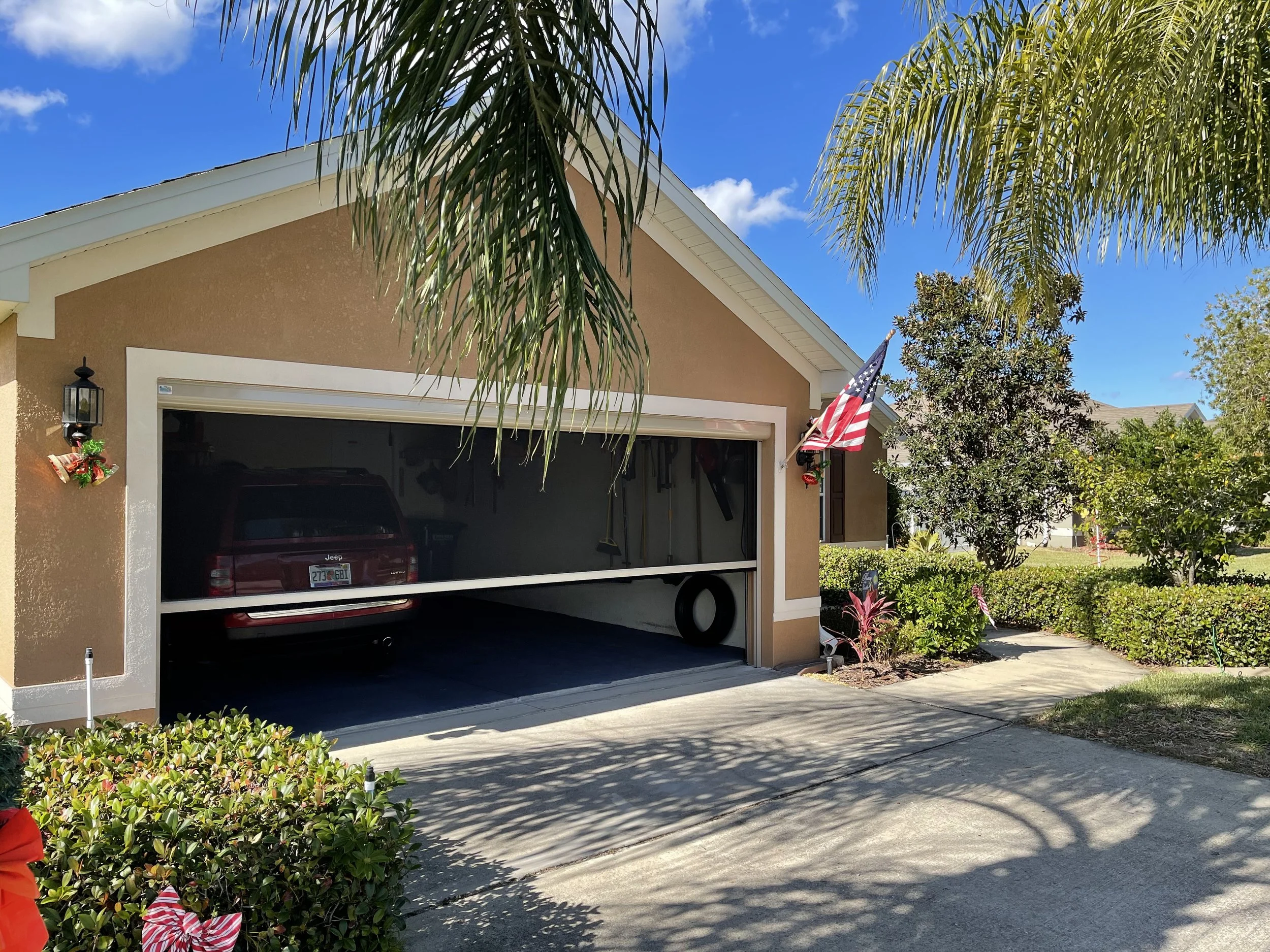 Front view of a garage attached to a house with holiday decorations, an American flag, and a red SUV inside. The driveway is partially shaded, with lush green bushes and tropical plants around. The sky is blue with some clouds.